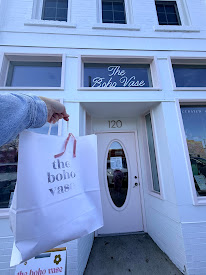 Person holding shopping bag in front of a white building with a sign reading 'The Boho Vase' above the door.