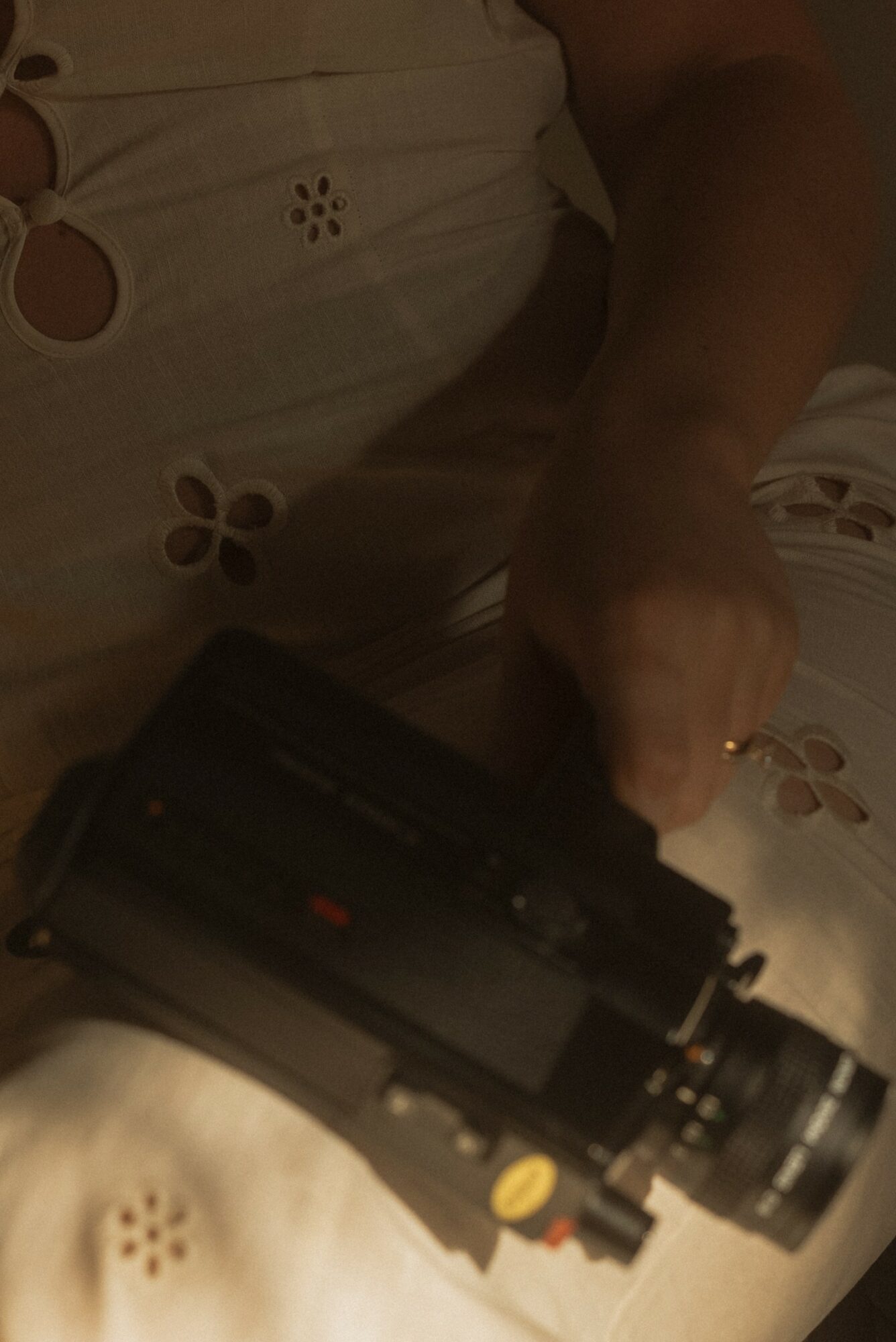 Person holding a black camera or device, wearing a white top with floral cutouts, in a dimly lit setting.