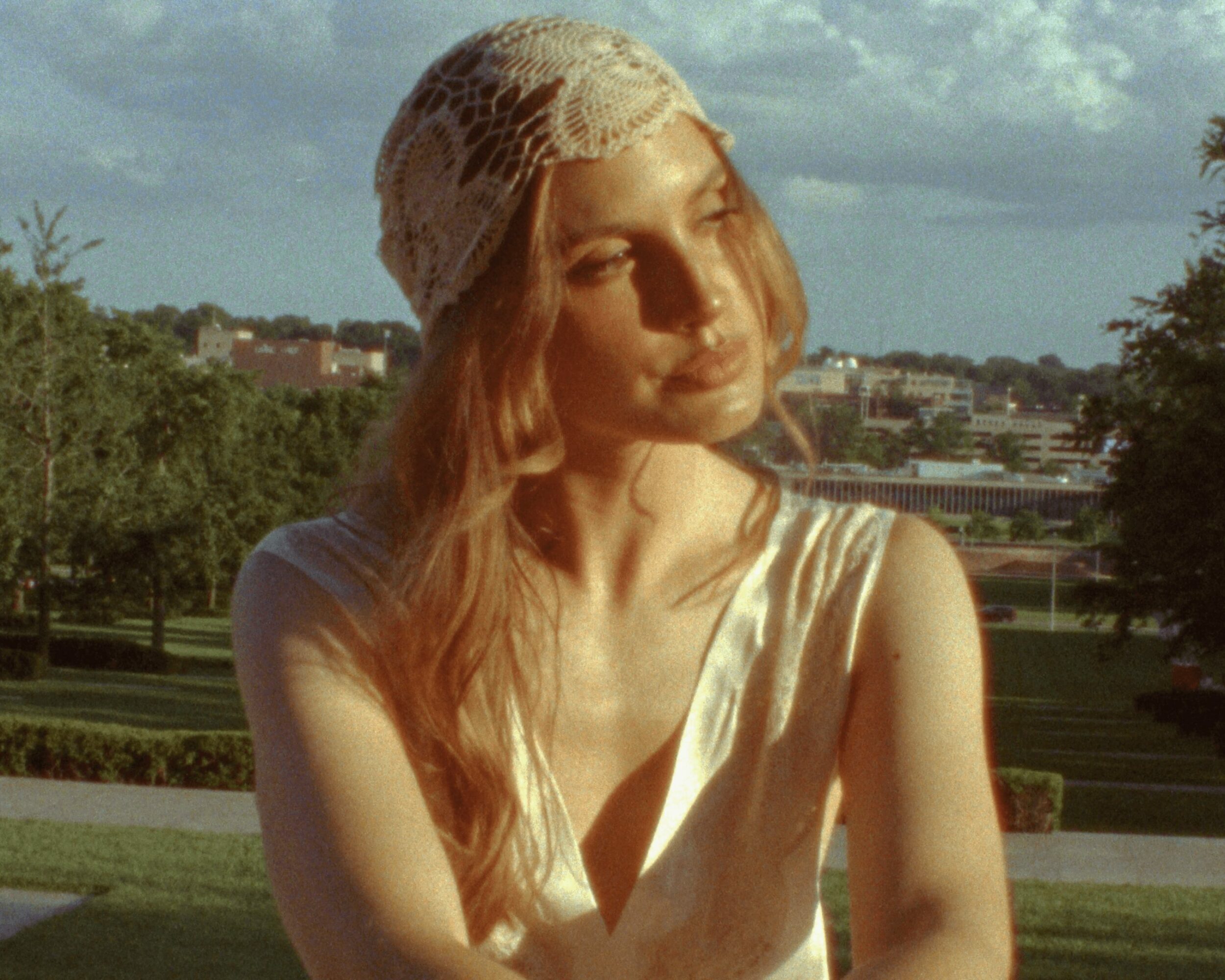 Young woman with long hair wearing a lace head covering, outdoors with trees and buildings in background.
