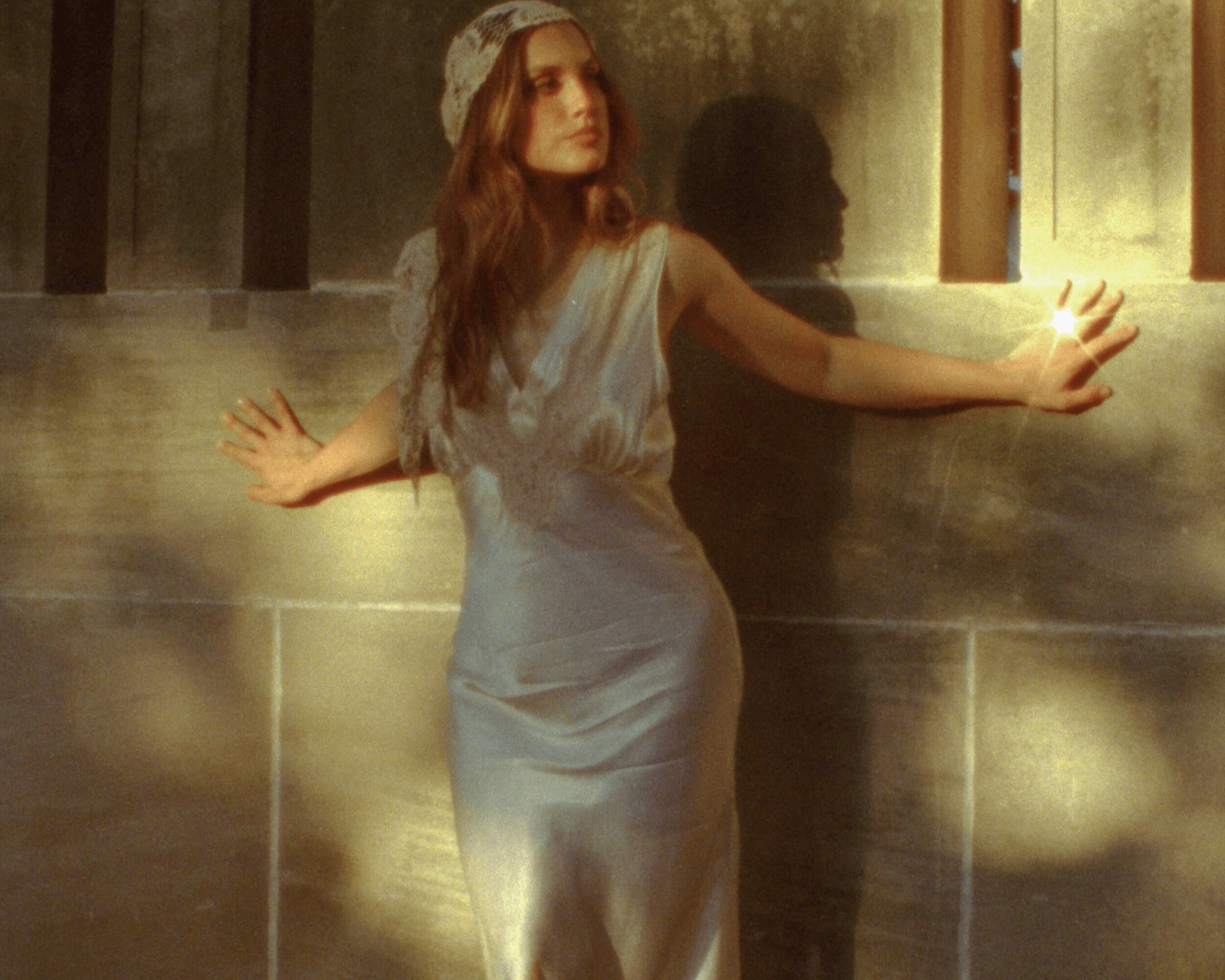 Woman in a light dress with arms outstretched, standing against a wall with sunlight and shadows.