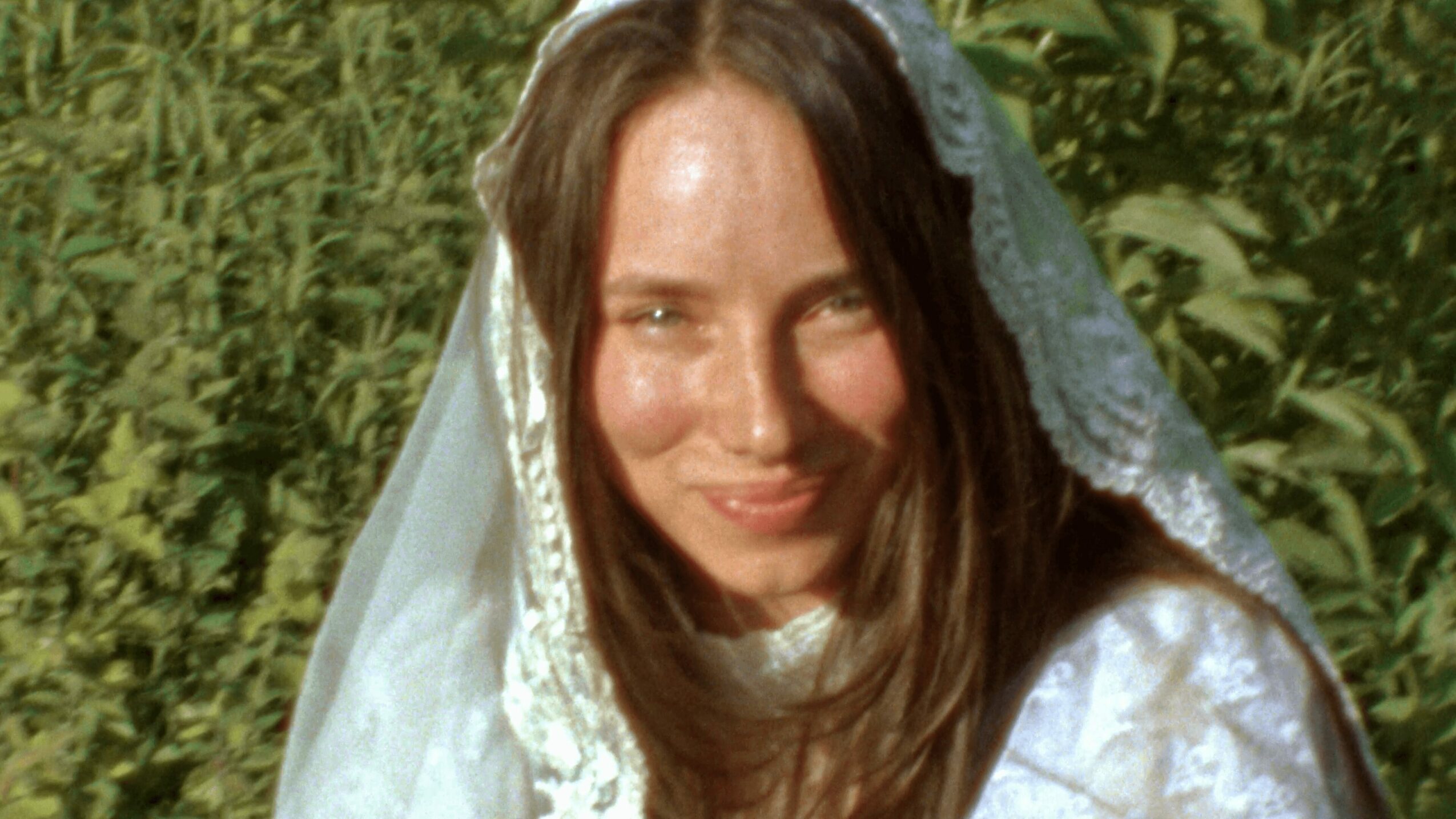 Young woman with long brown hair wearing a lace veil outdoors, smiling, surrounded by greenery.