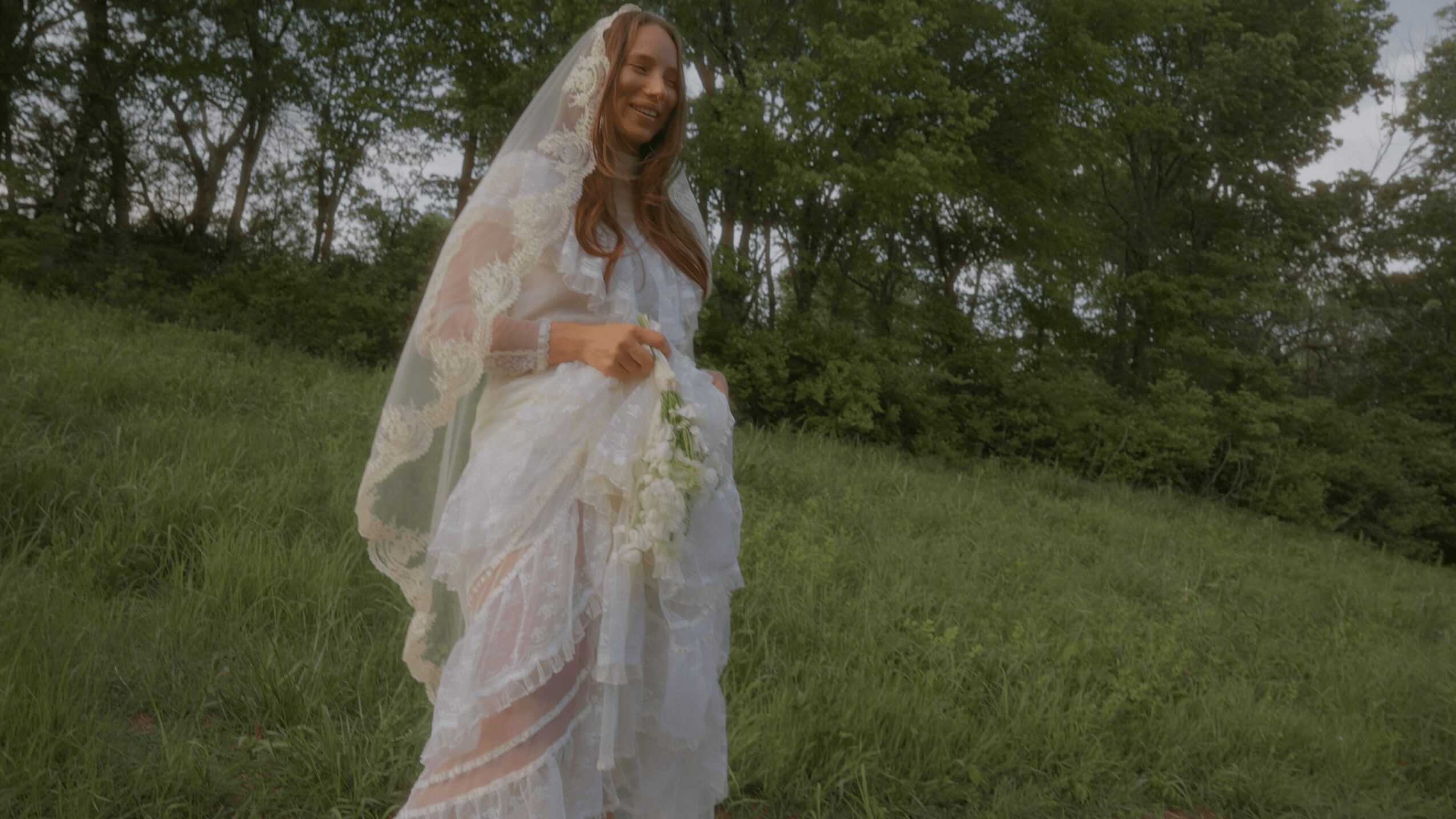Woman in wedding dress and veil standing outdoors on grass with trees in background.