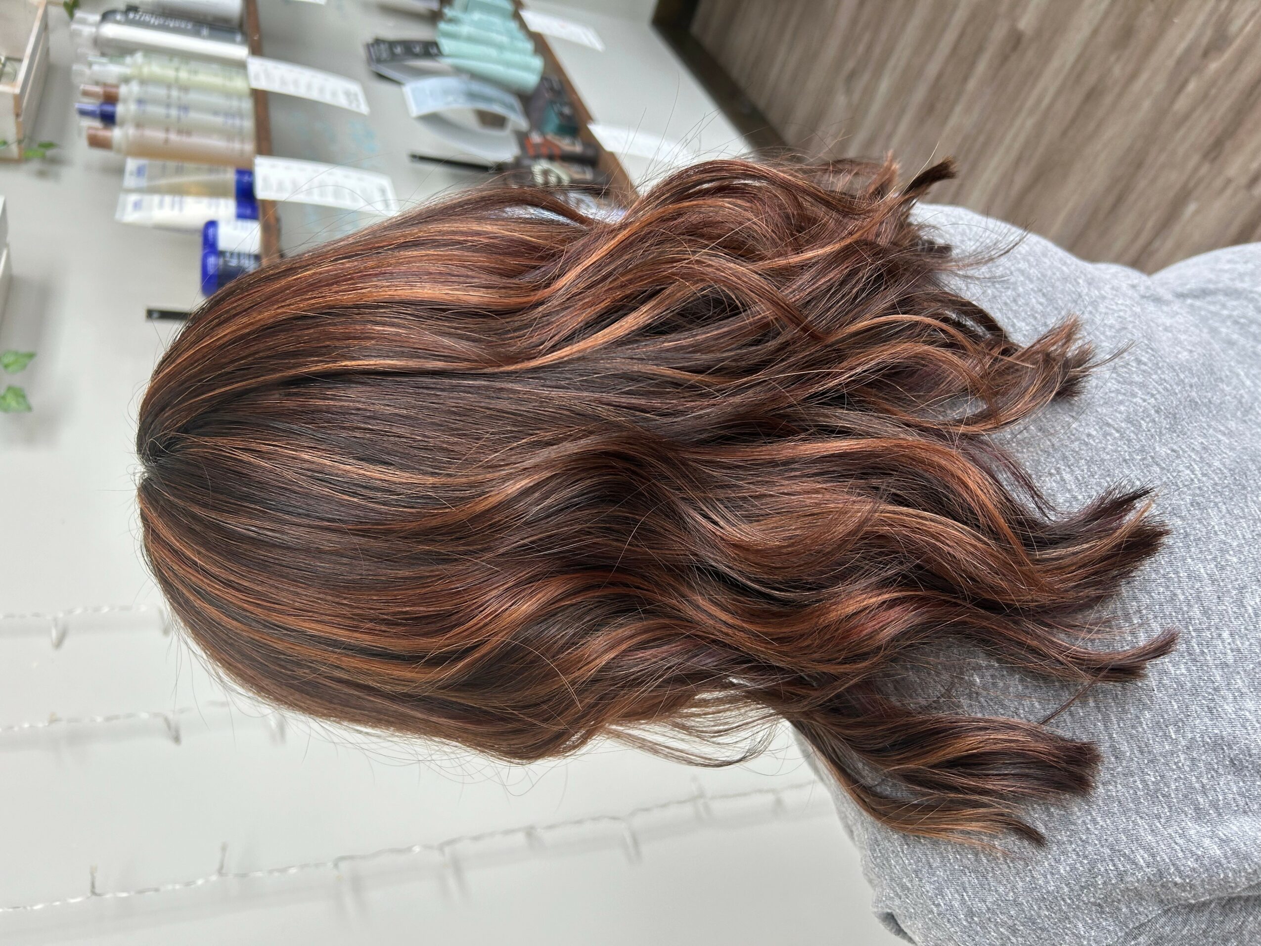 Side view of a woman with wavy, reddish-brown hair, in a salon setting with hair products in the background.