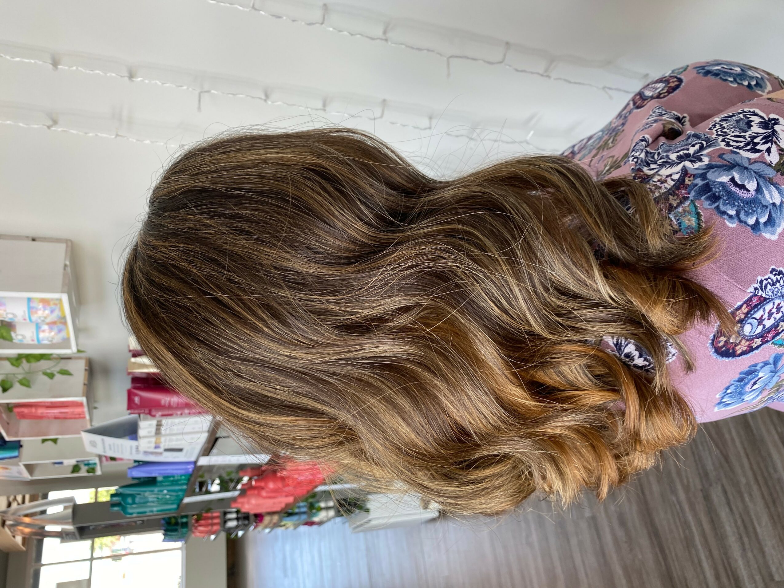Person with wavy, shoulder-length hair facing away, in a room with shelves and books, wearing a floral top.