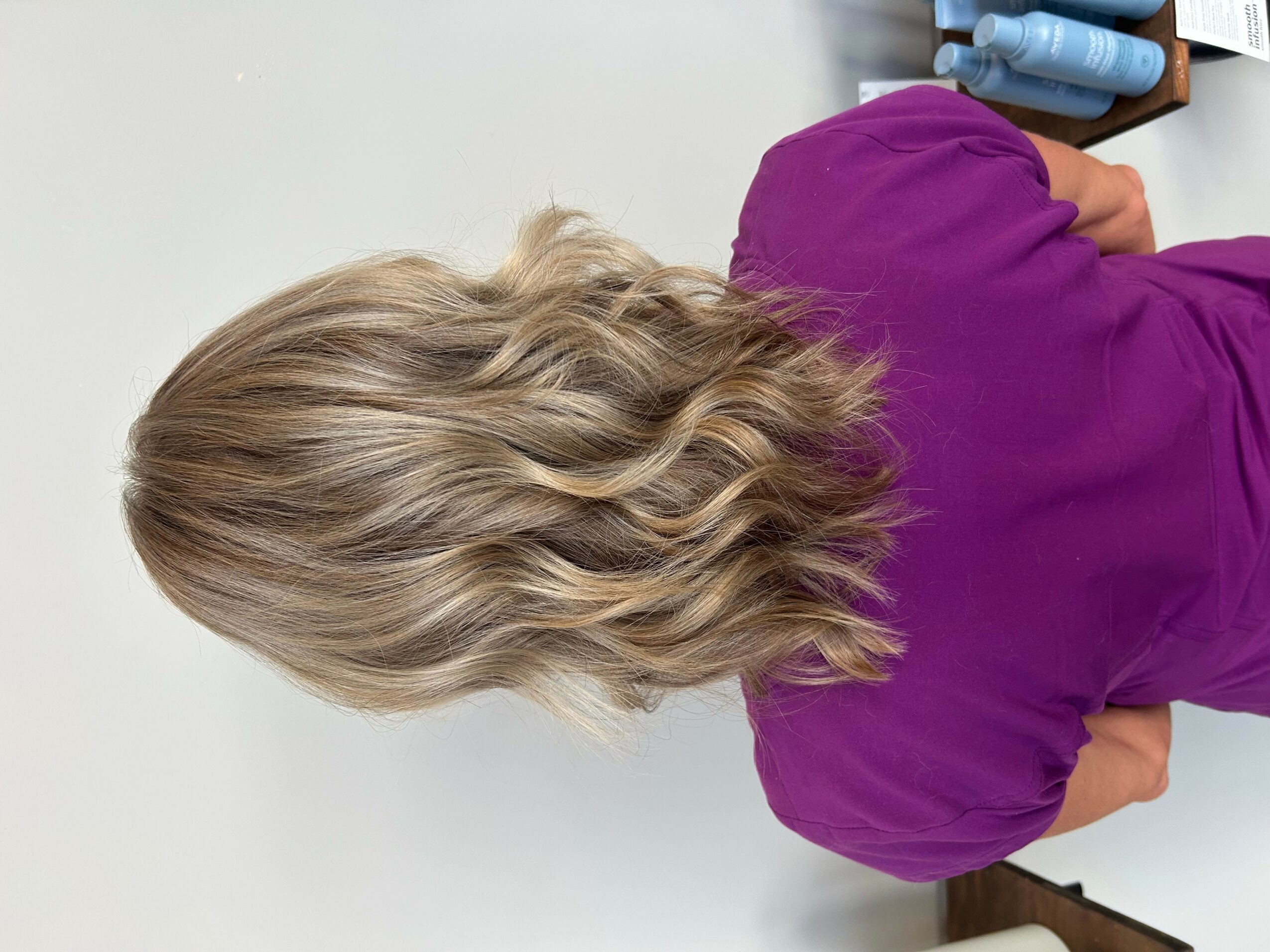 Person with wavy blonde hair wearing a purple shirt, viewed from behind, sitting at a table with bottles.