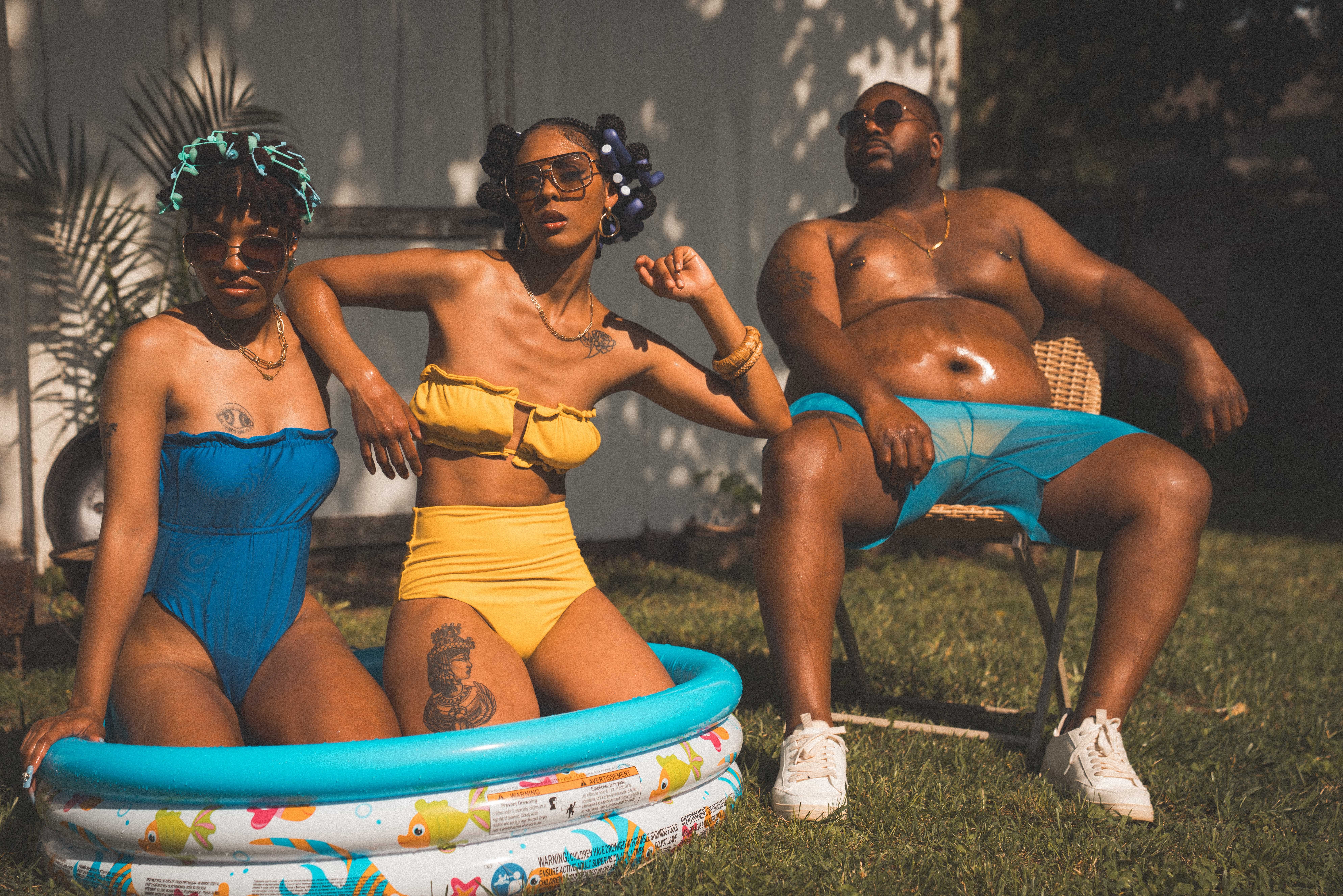 Three people outdoors, two women in swimsuits and a man in shorts, sitting and relaxing on chairs and a pool.