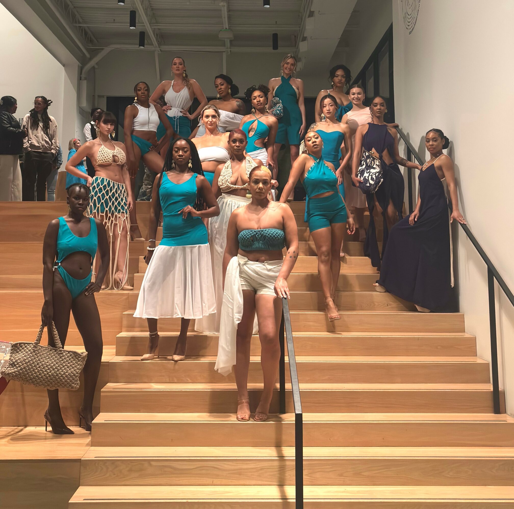 Group of women on wooden stairs in colorful dresses, some posing, in an indoor setting with white walls.