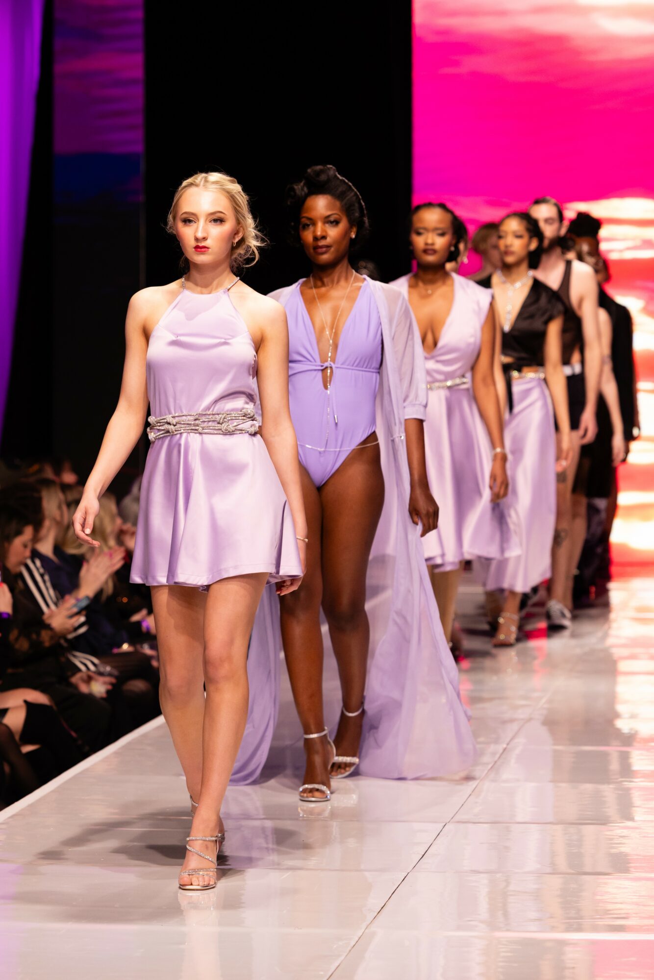 Group of models walking down runway in pastel purple and lavender dresses, with a pink and purple background.