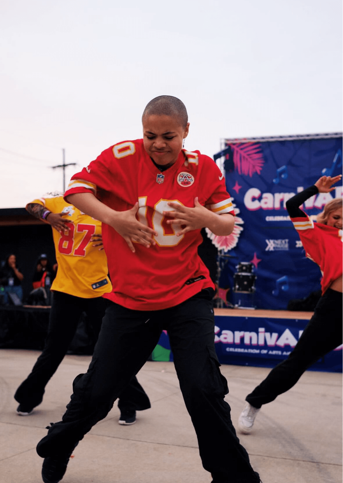 Child dancing outdoors, wearing a red sports jersey, with other children and a carnival banner in the background.