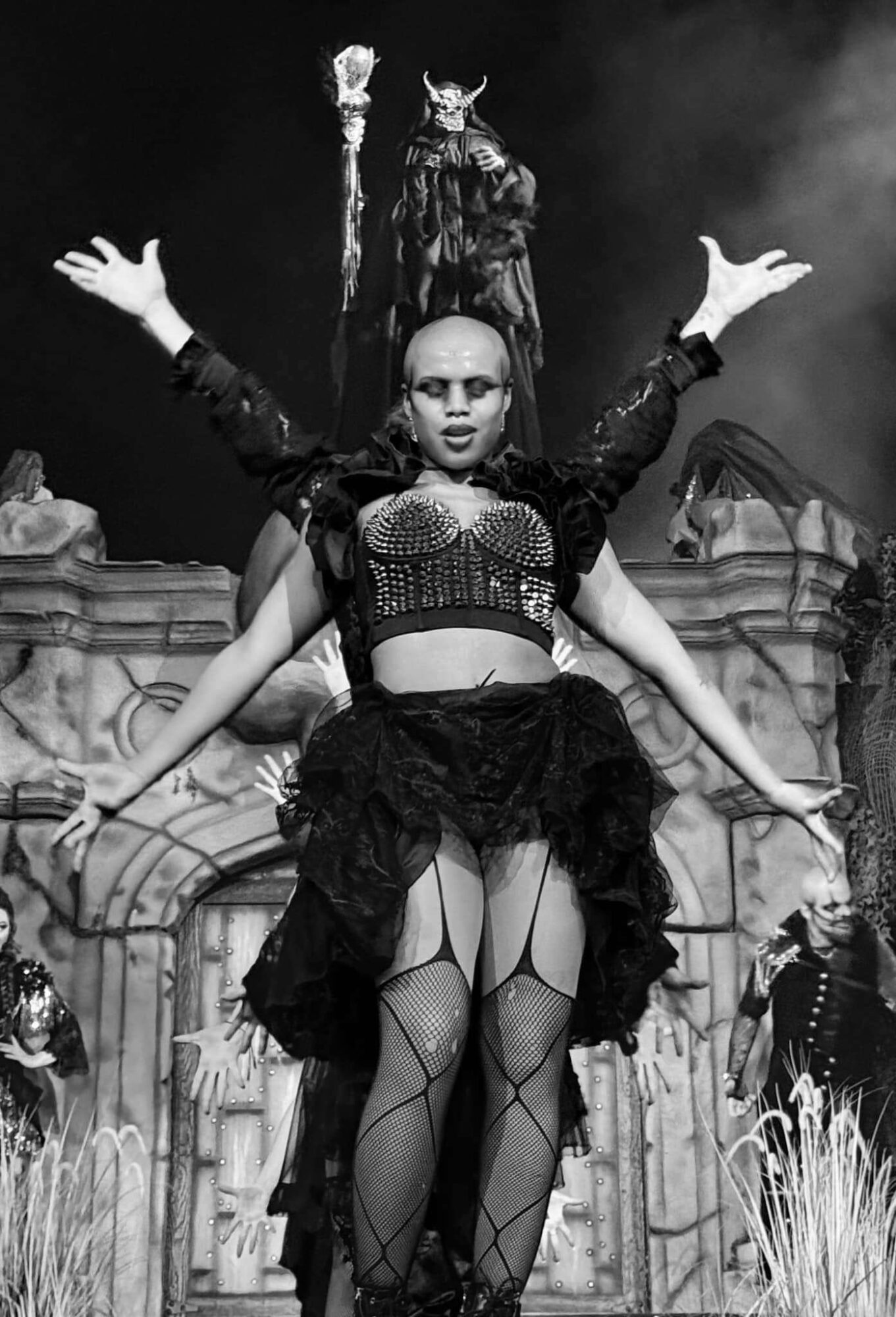 Group of performers on stage with a central figure in dark clothing and makeup, arms raised, in front of a gothic backdrop.