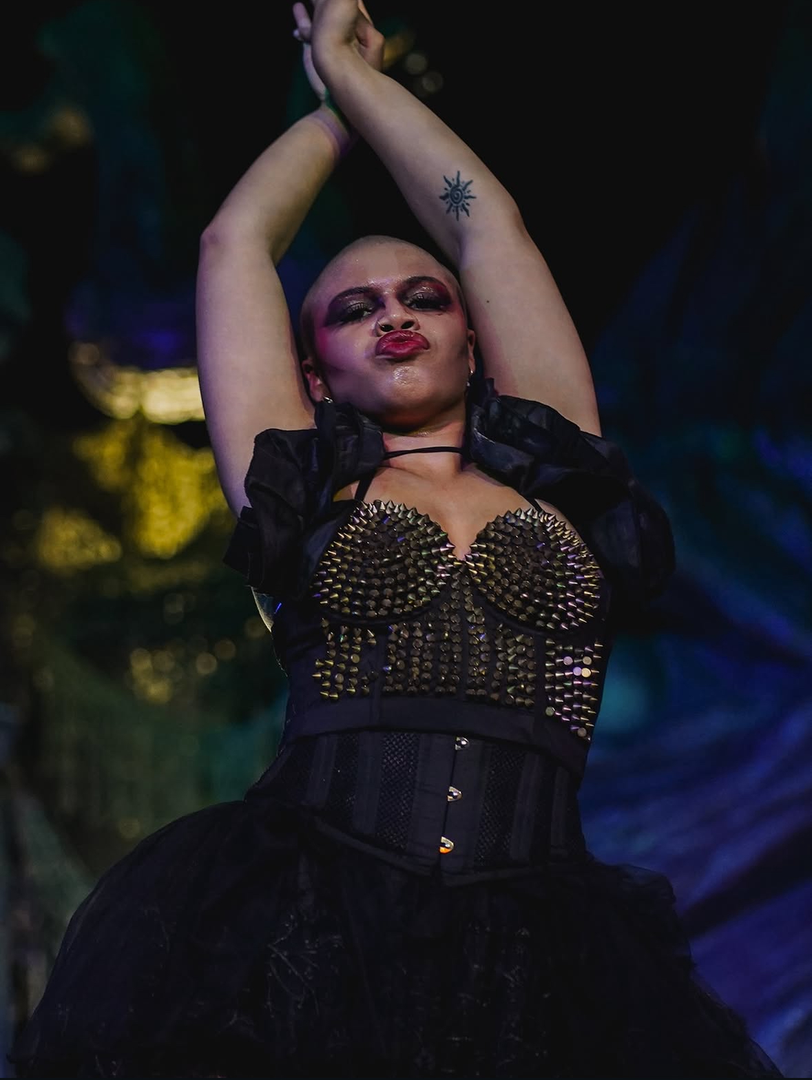 Person with arms raised, wearing dark clothing and makeup, with a tattoo on arm, in a dark, colorful background.