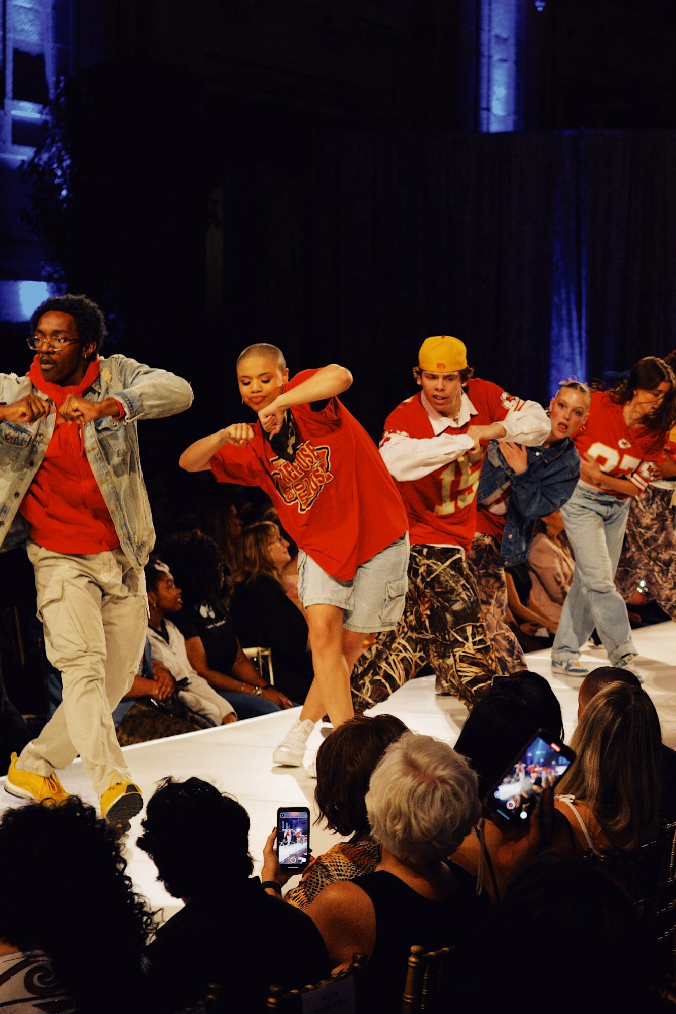 Group of children dancing on stage with audience watching, some taking photos, in a dark setting with blue lighting.