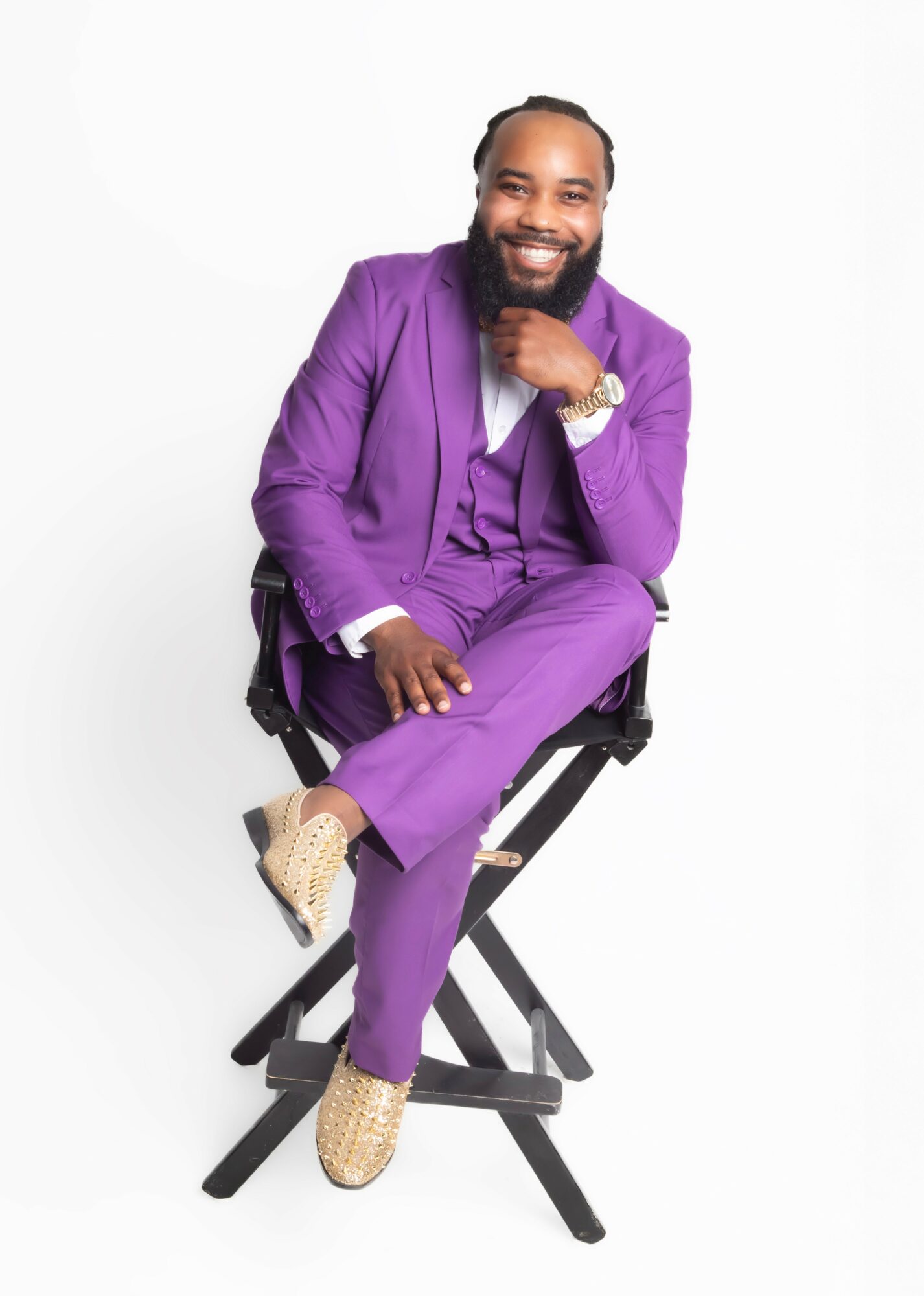 Man with a beard in a purple suit sitting on a black chair, smiling, with a white background.