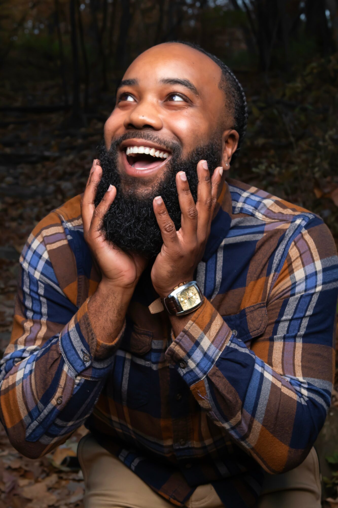 Man with a beard smiling and touching his face outdoors in a wooded area, wearing a plaid shirt and watch.