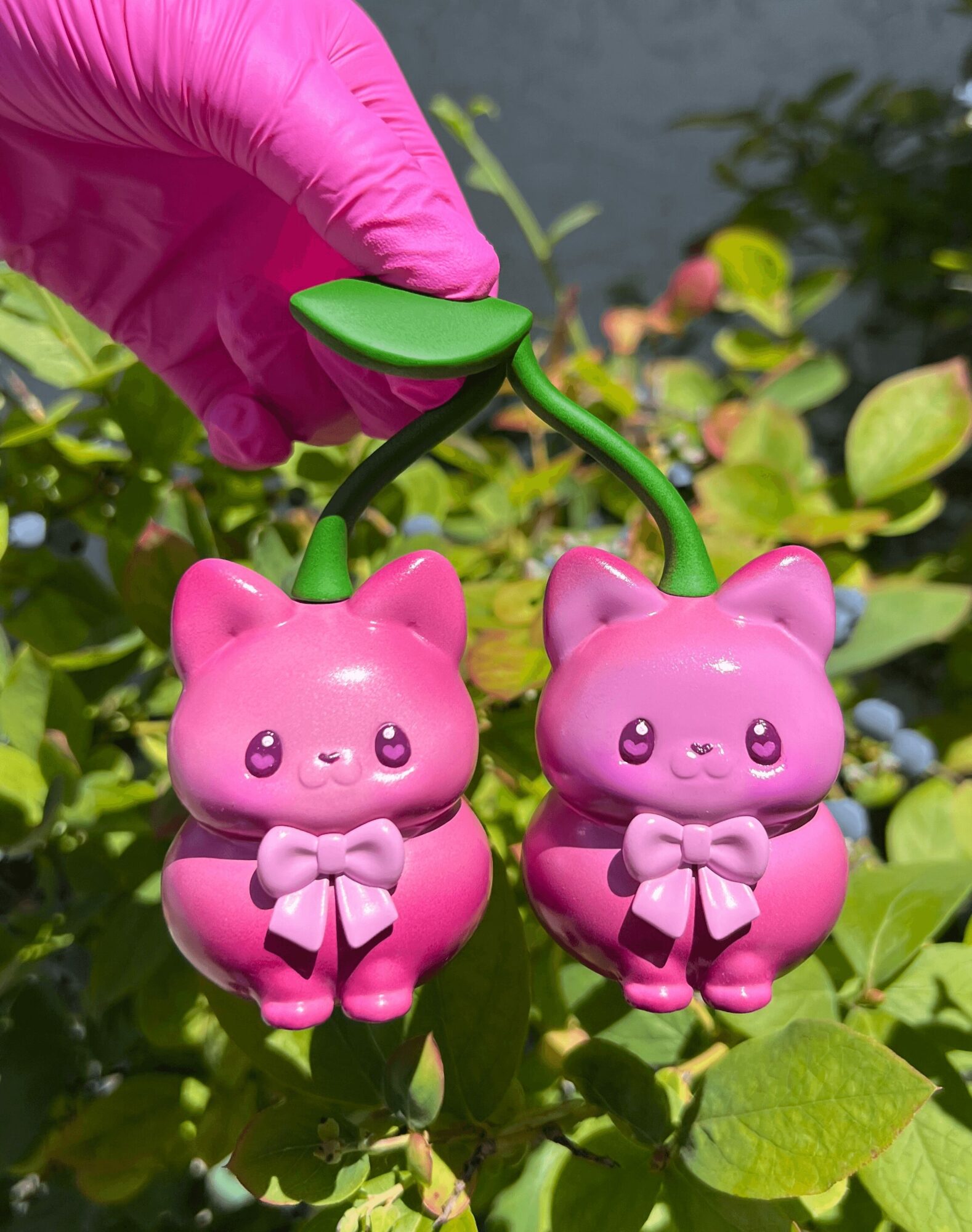 Pink cat-shaped keychain with bows, held by a hand in a pink glove, hanging among green leaves.