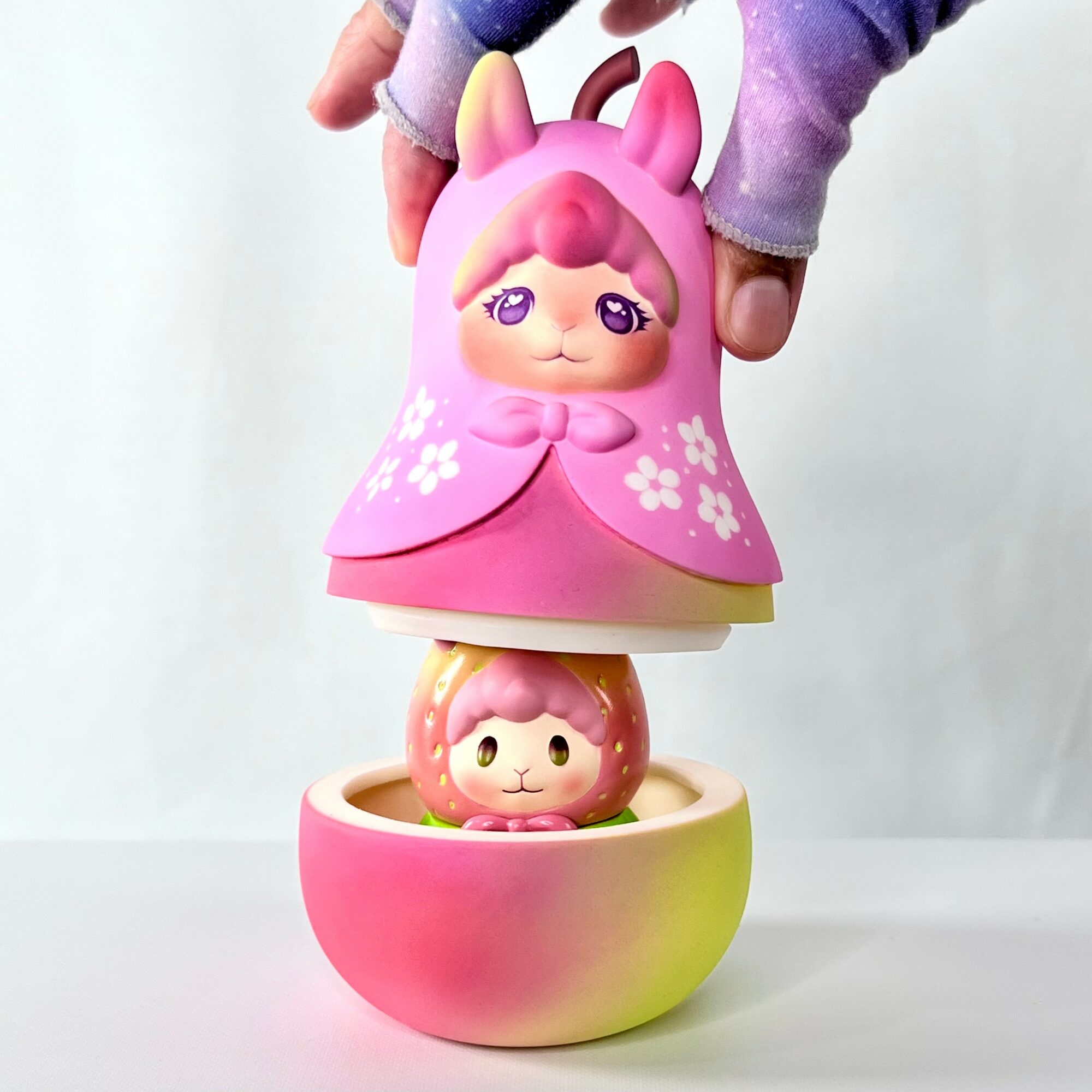 Person holding a pink unicorn-themed toy with a doll inside a pink and yellow container, against a plain background.
