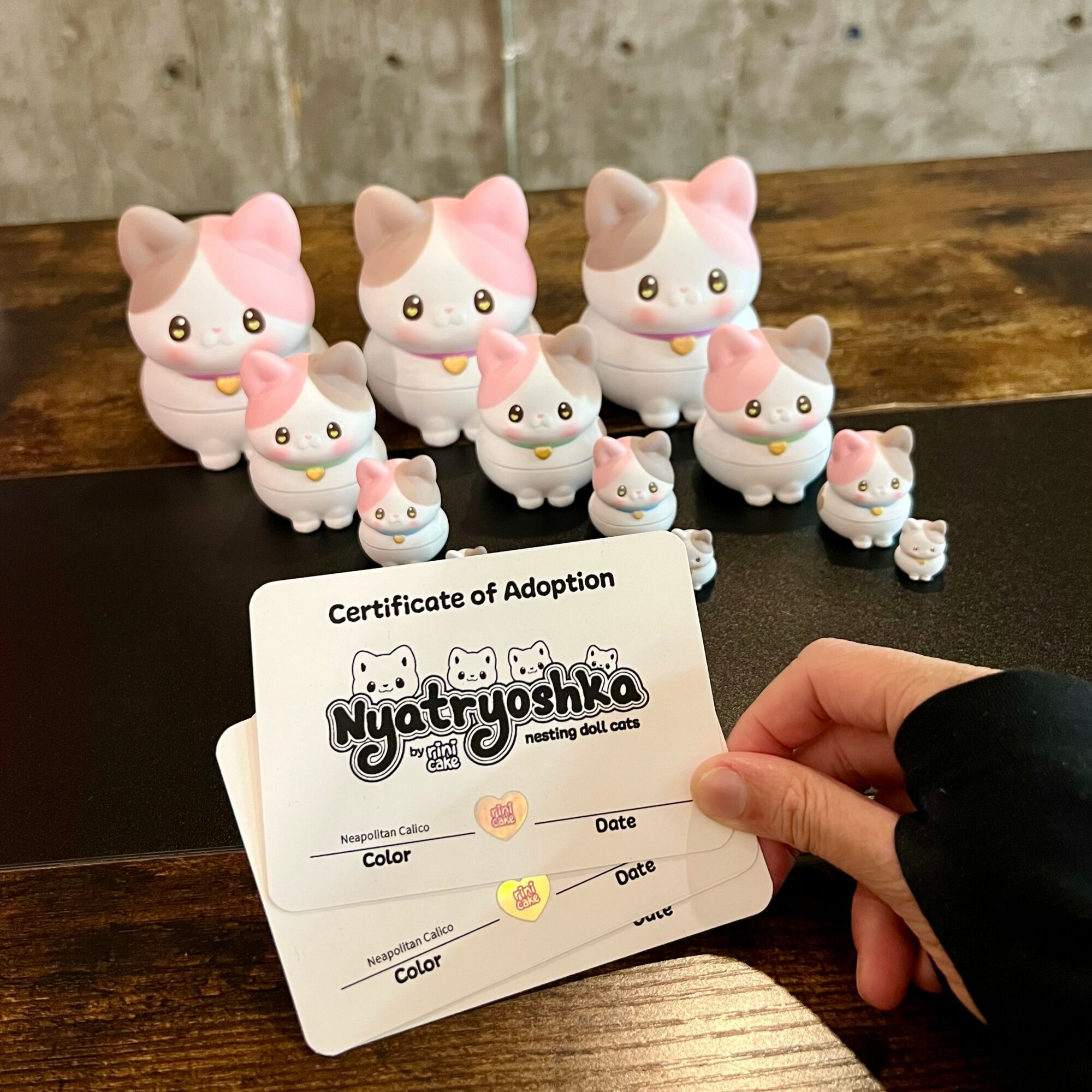 Collection of nine small pink and white cat figurines with a hand holding a certificate of adoption in front.