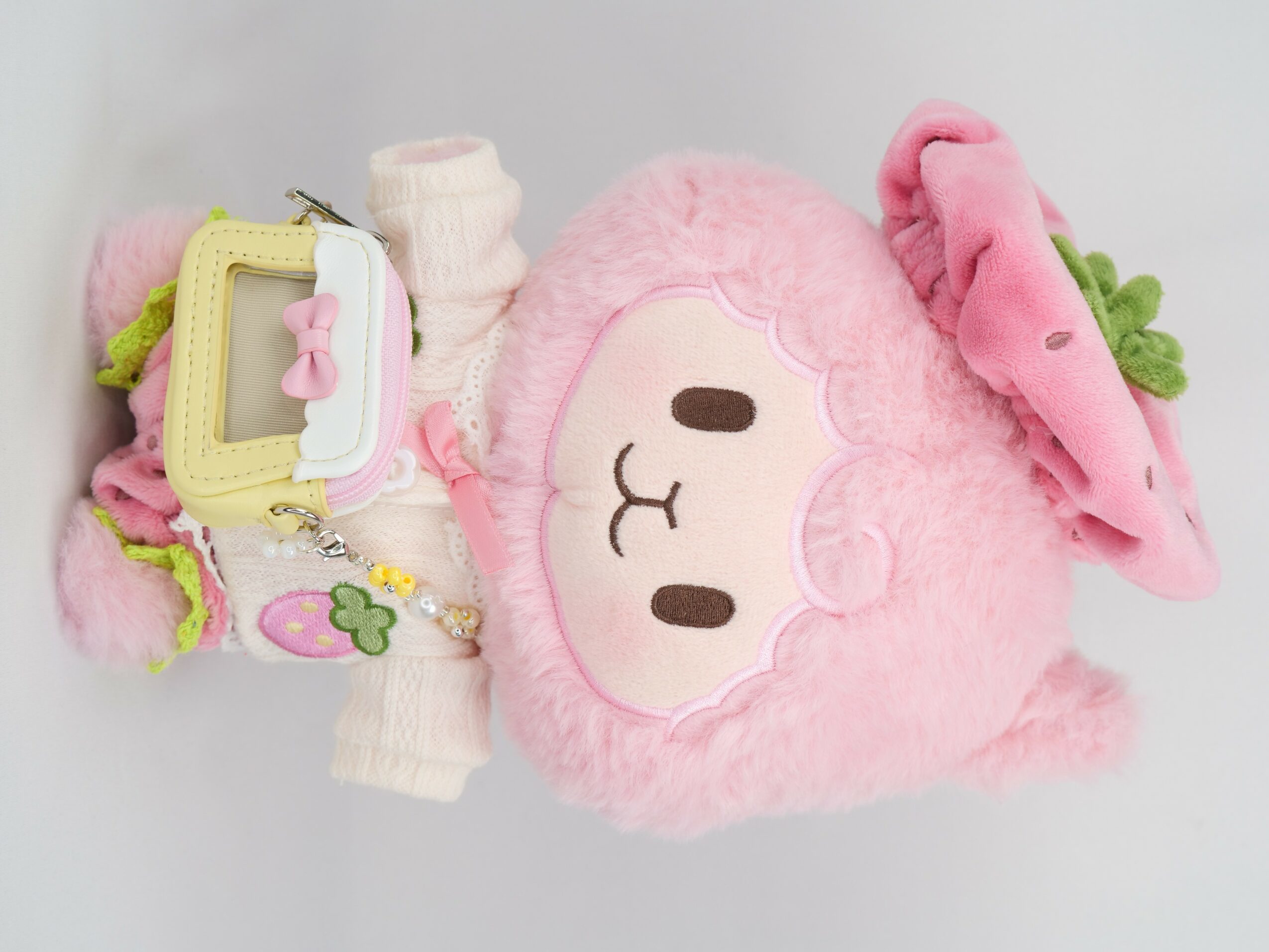 Pink plush toy with a face, wearing a pink hat with green leaves, and a colorful wristband with charms.