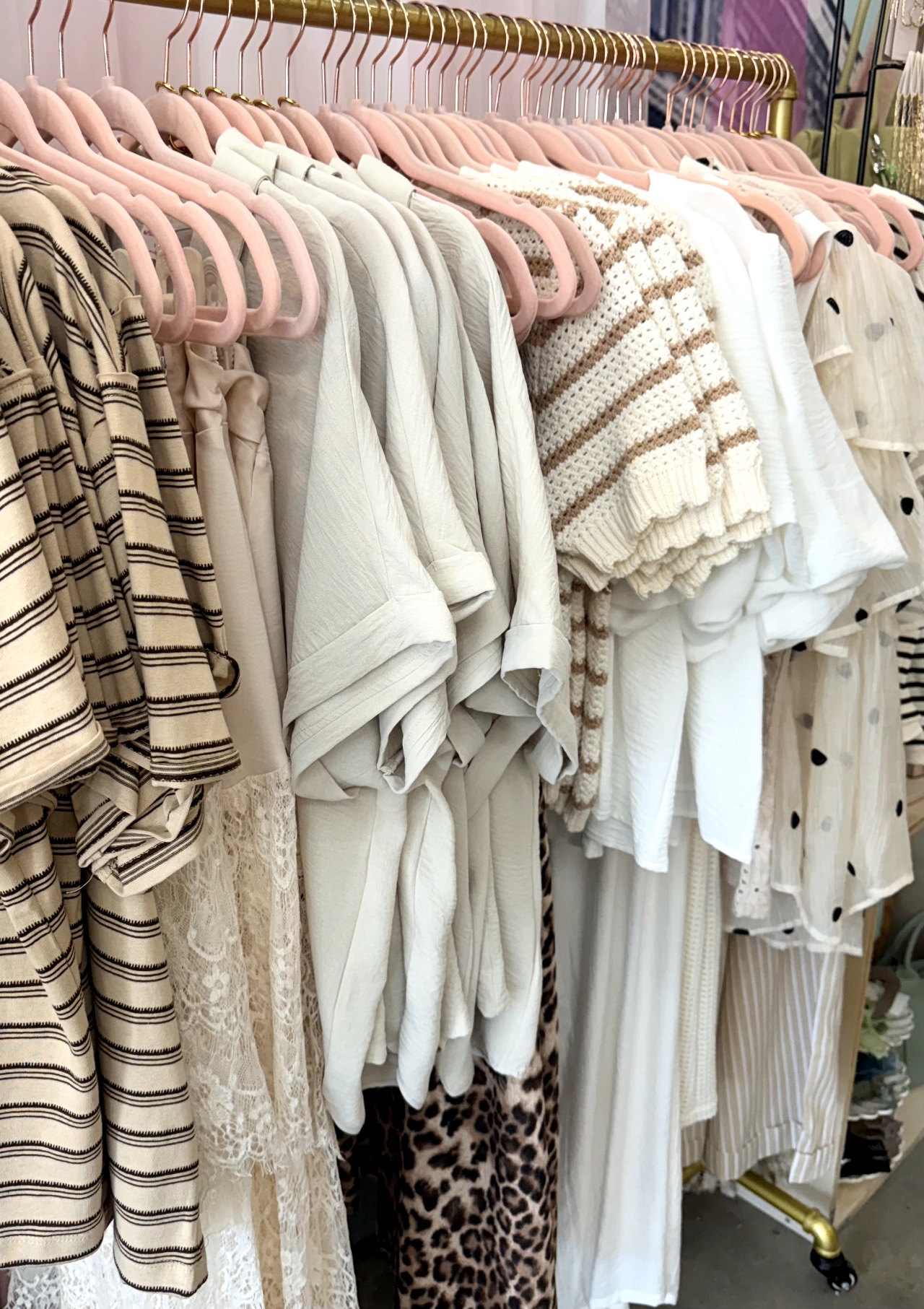 Clothing racks with various light-colored and patterned garments, including sweaters, dresses, and shirts, hanging on pink hangers.