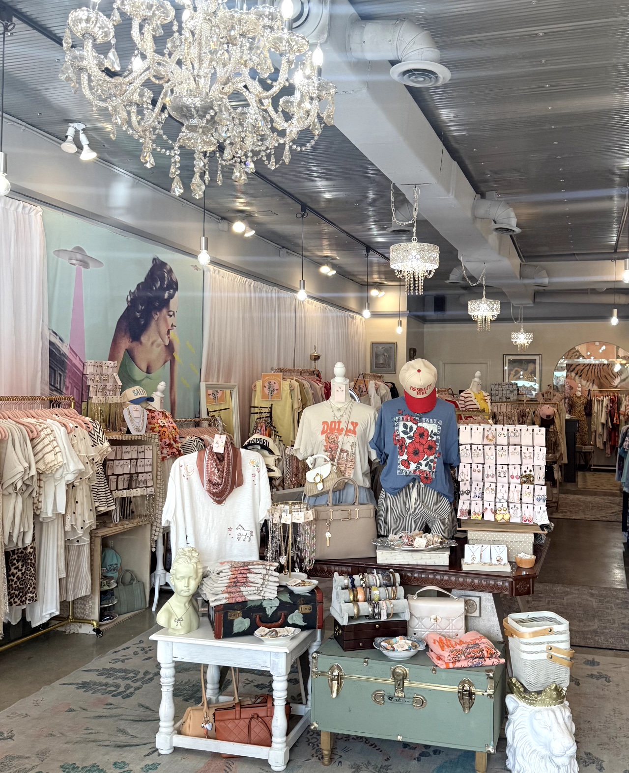 Clothing store interior with mannequins, racks of clothes, chandeliers, and decorative lighting, displaying various apparel and accessories.
