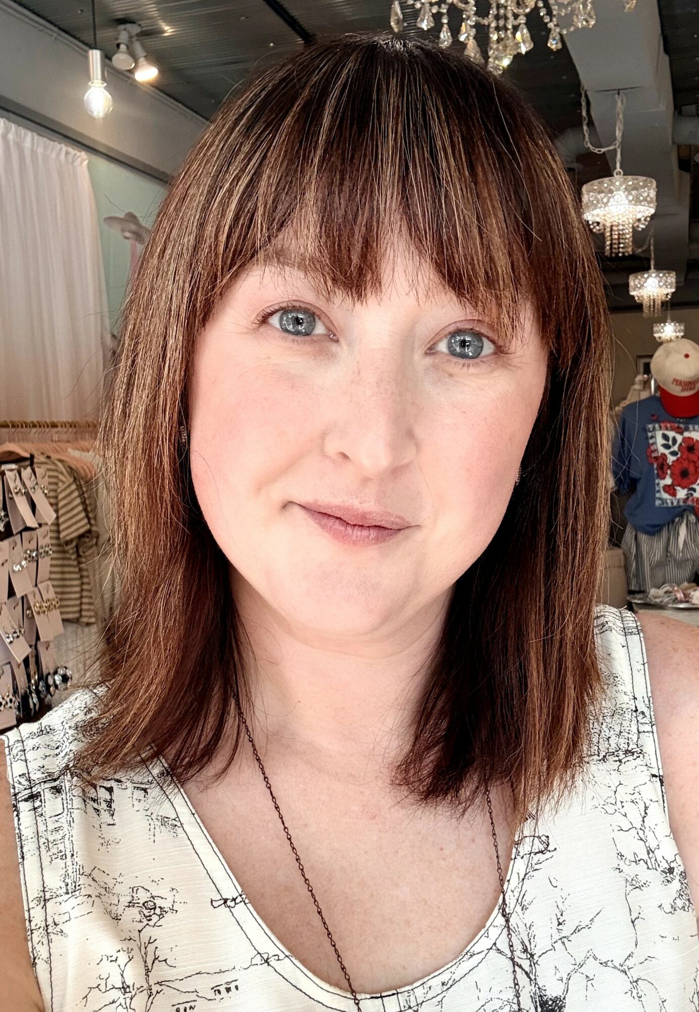 Woman with shoulder-length brown hair and bangs, smiling, wearing a white patterned top, in a store with chandeliers and mannequins.
