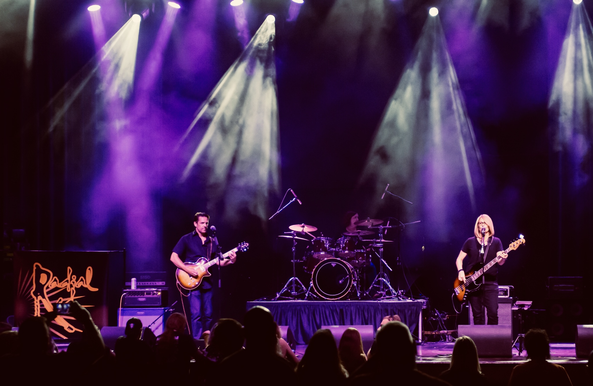Band performs on stage with purple and white lighting, audience silhouettes visible, musical instruments and equipment present.