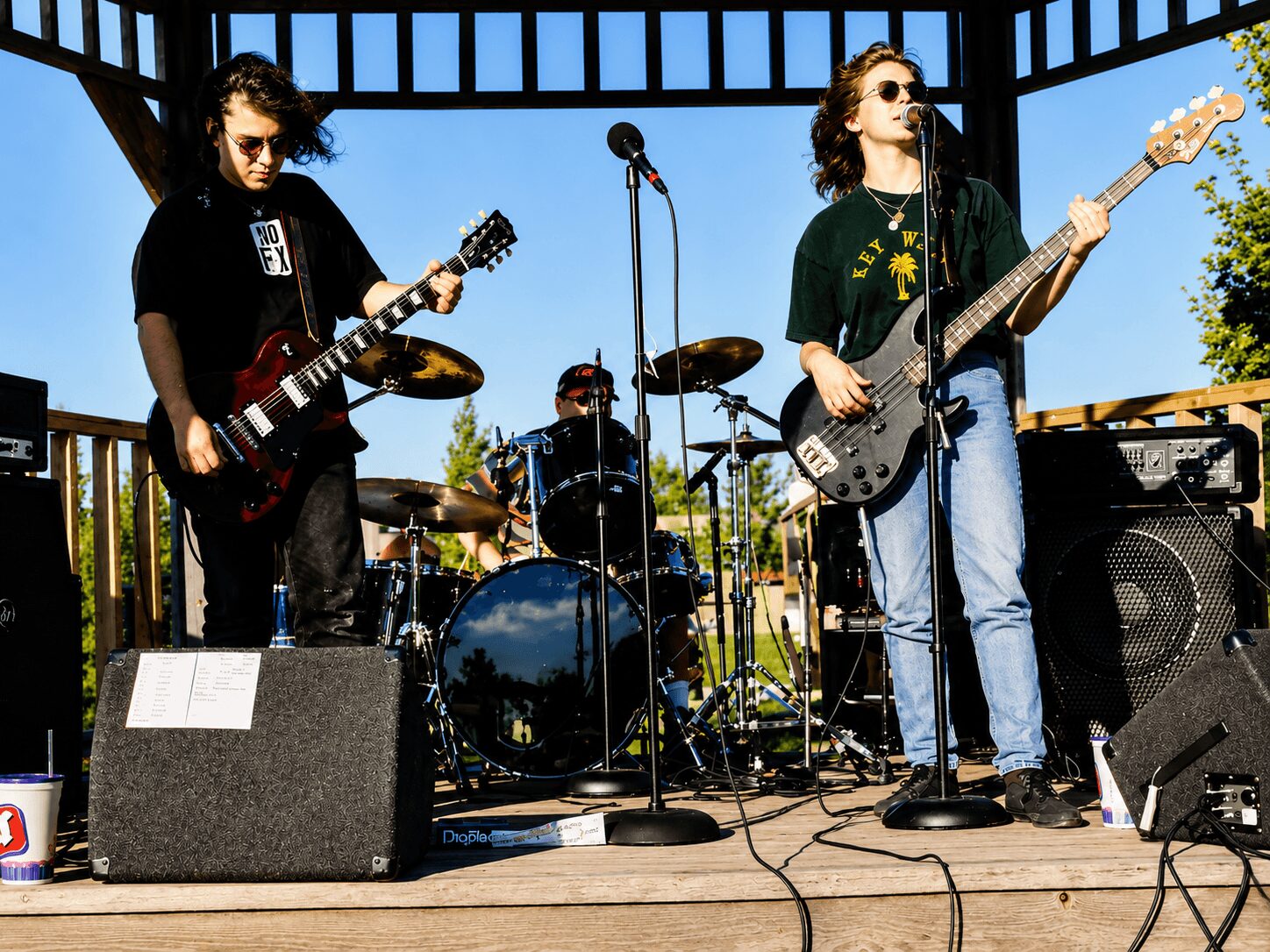 Three musicians perform on an outdoor stage with guitars and drums, under a wooden pergola, with trees in the background.