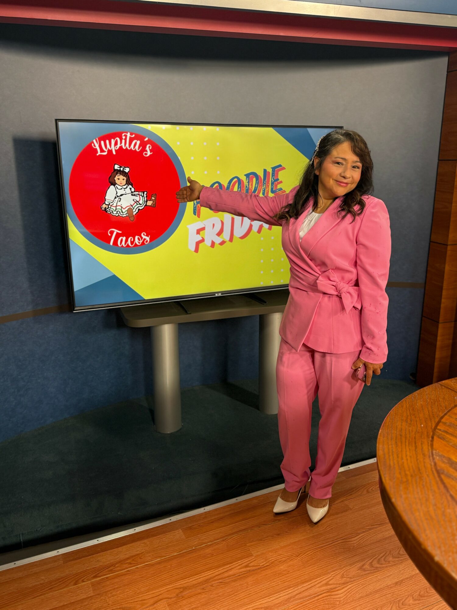 Woman in pink suit standing next to a screen with a colorful logo and text, smiling, in an indoor setting.