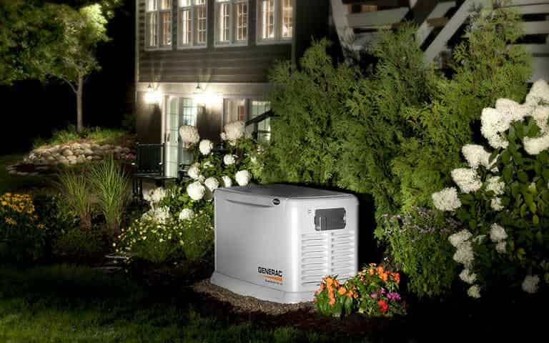 Nighttime garden scene with a white generator, flowers, bushes, and a house in the background.