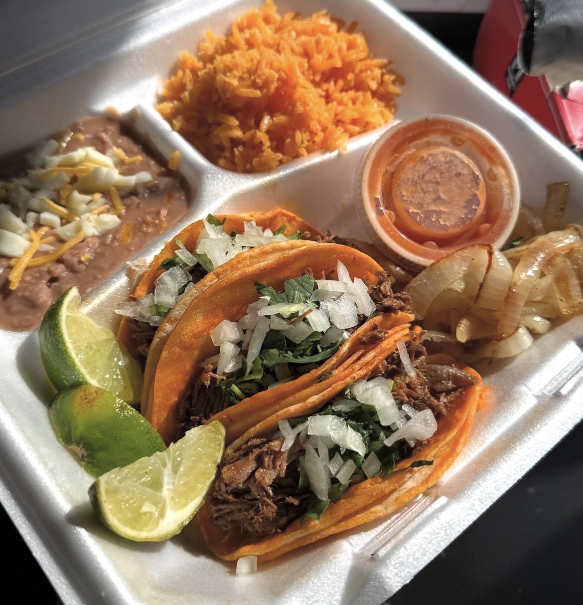 Mexican tacos with shredded meat, chopped onions, and cilantro, served with lime wedges, rice, and grilled onions in a foam tray.