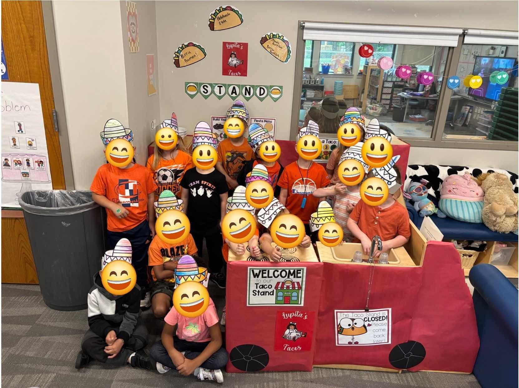 Group of children and adults in a classroom with a red display stand, smiling emojis covering faces, colorful decorations, and educational posters.