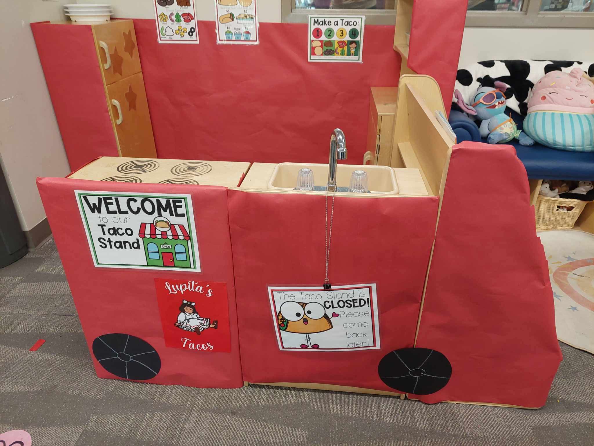 Red play kitchen with a sink, faucet, and signs including a welcome message and a closed sign with a dog, on a carpeted floor.