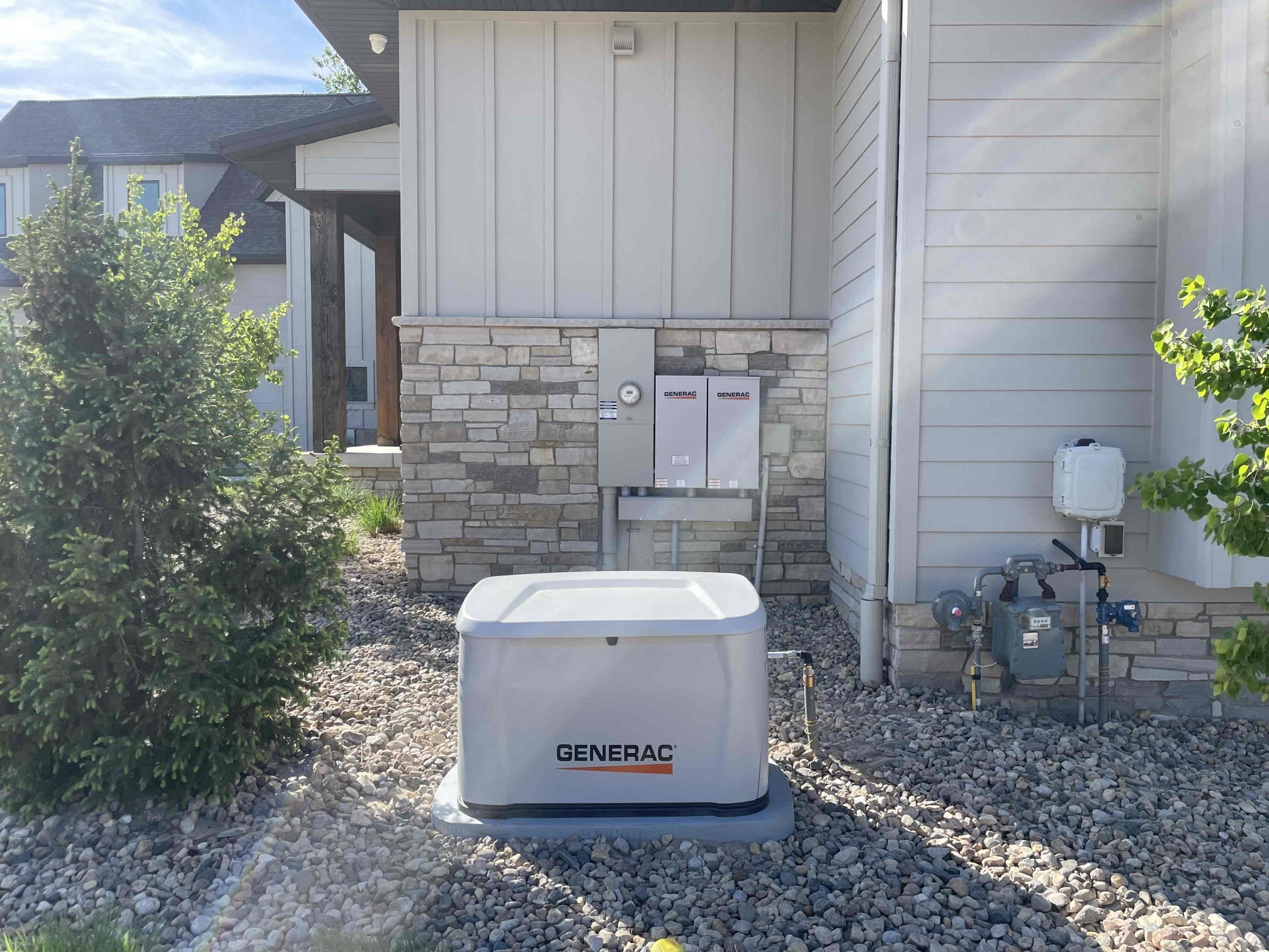 Outdoor generator with electrical boxes on a wall, gravel ground, and a bush on the left.