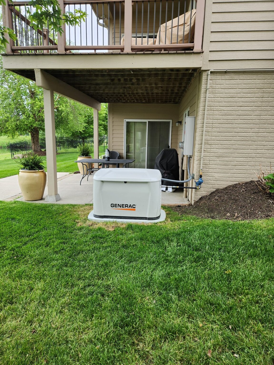 Backyard with a generator, patio furniture, and potted plant under a balcony, with green grass and trees in the background.