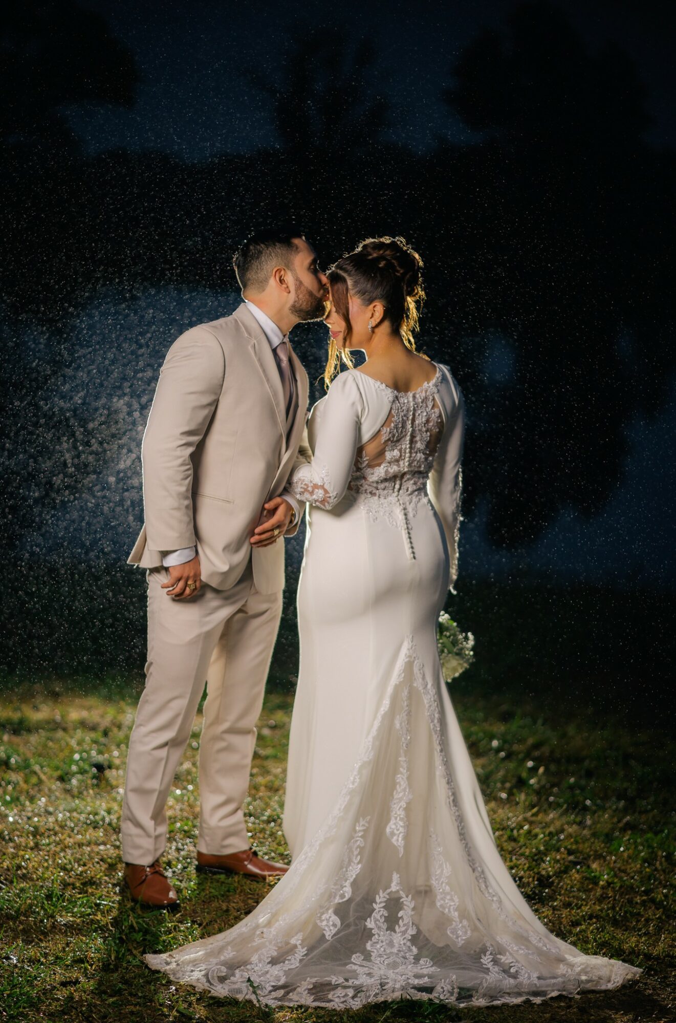 Bride and groom stand close outdoors at night, holding hands, with water droplets falling around them, illuminated softly.
