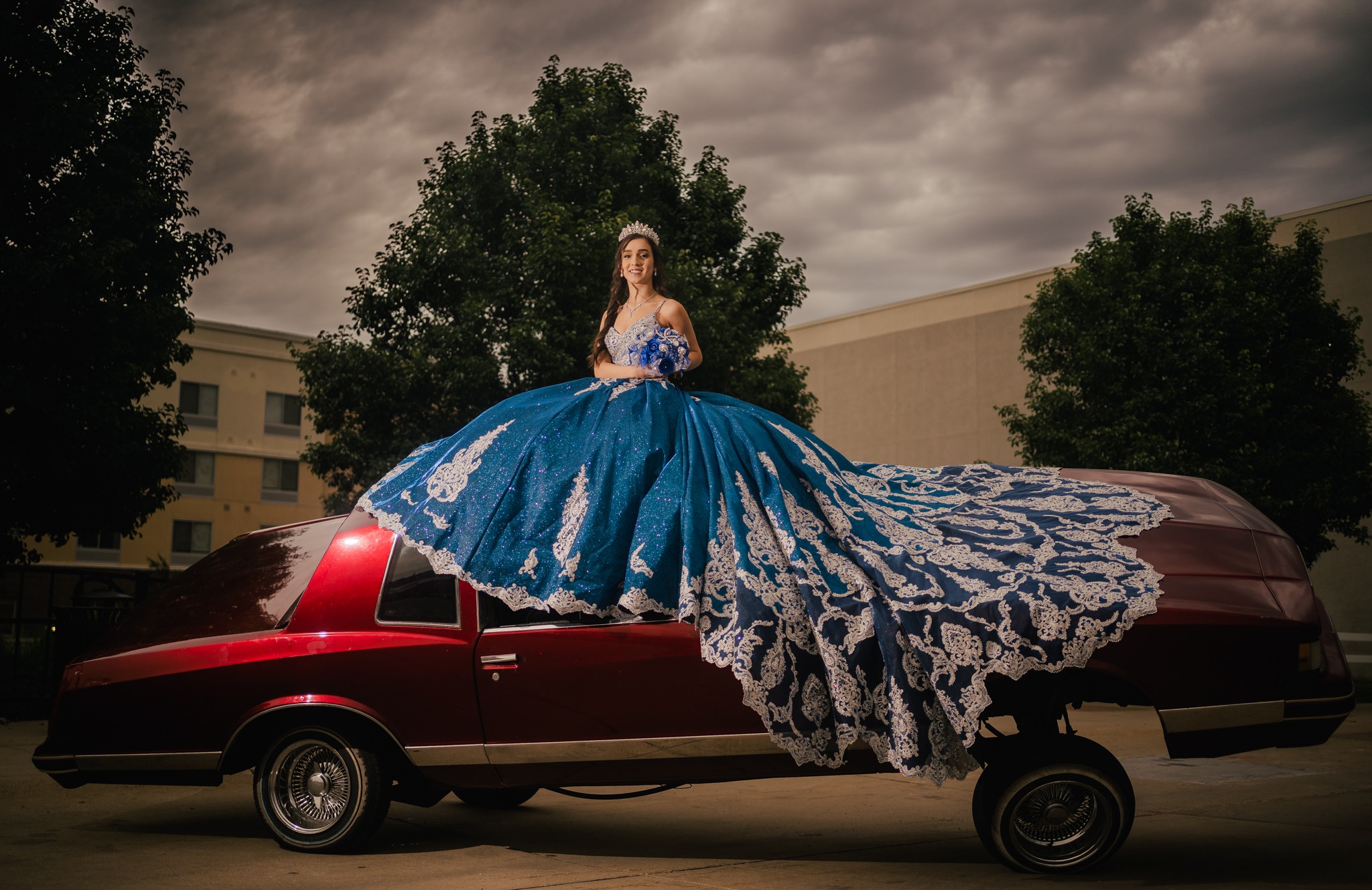 A woman in a blue and white gown stands on a red limousine, with trees and buildings in the background.