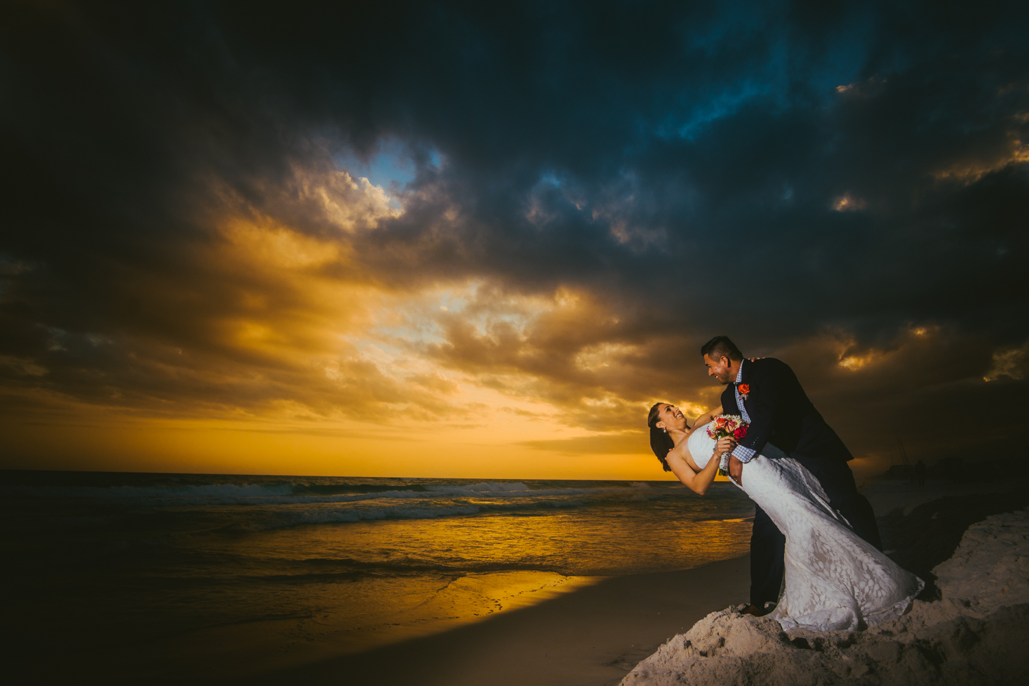 A couple dancing on a beach at sunset with dramatic clouds and colorful sky in the background.