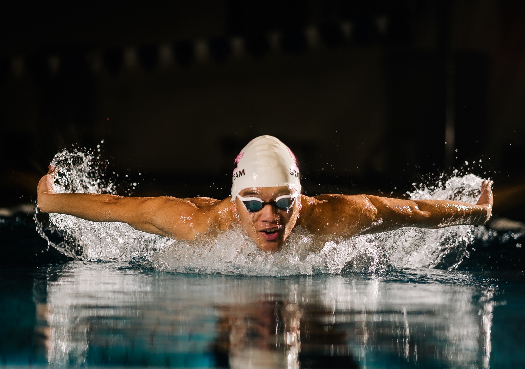 Swimmer in a pool performing a butterfly stroke with arms extended and water splashing around.