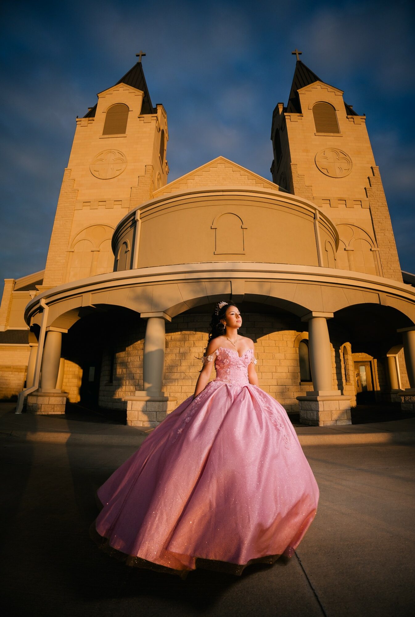 Woman in pink gown standing in front of a church with twin towers, under a cloudy sky.