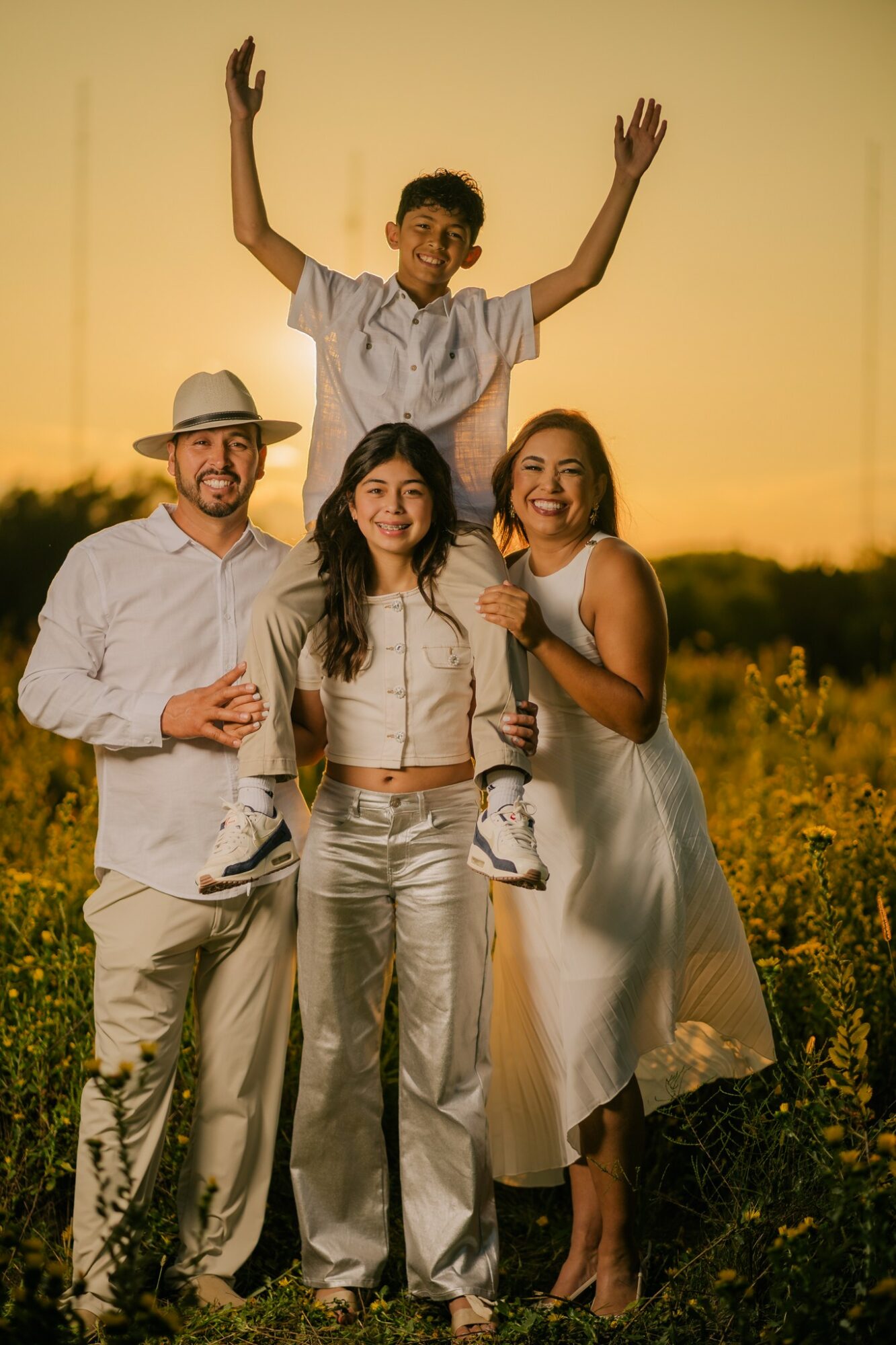 Group of four people outdoors during sunset, smiling, with one person standing on others' shoulders, arms raised.