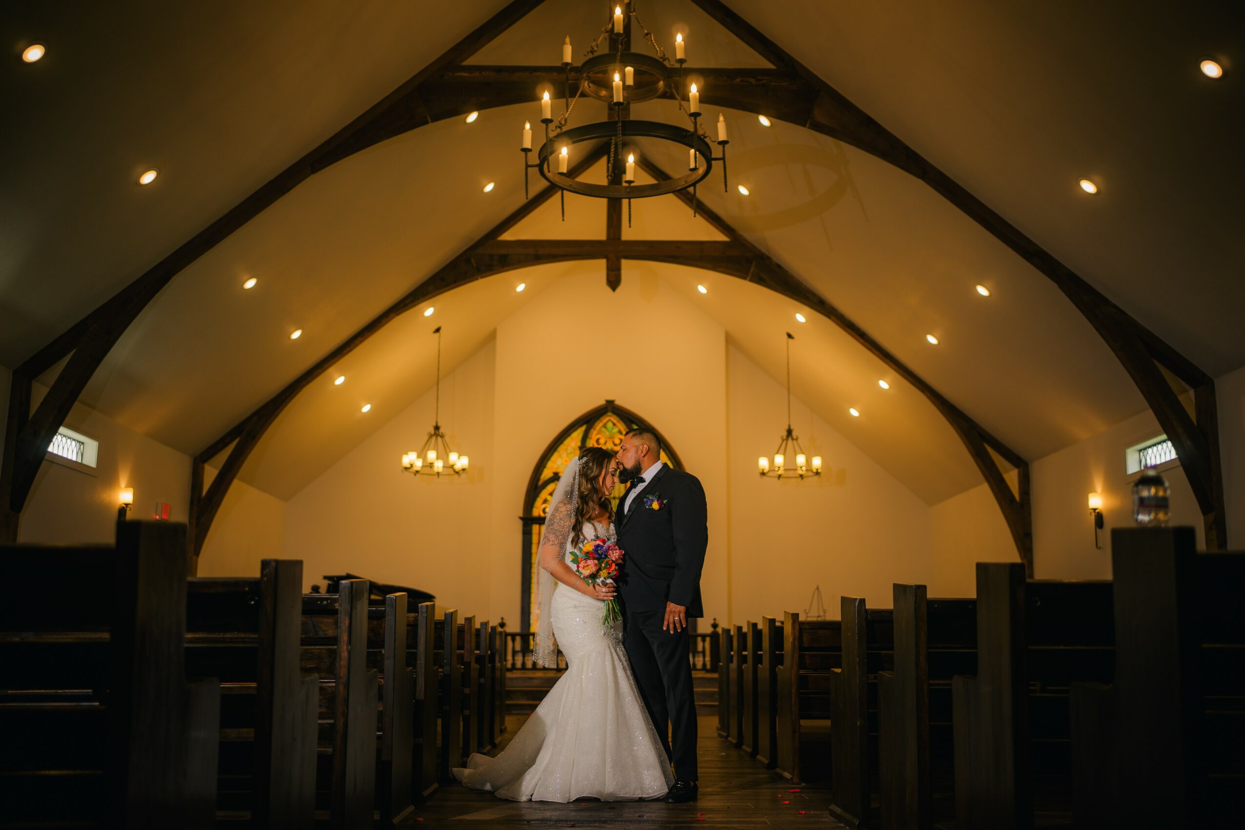 A bride and groom stand close together in a church aisle, holding hands and facing each other, with empty pews around them.