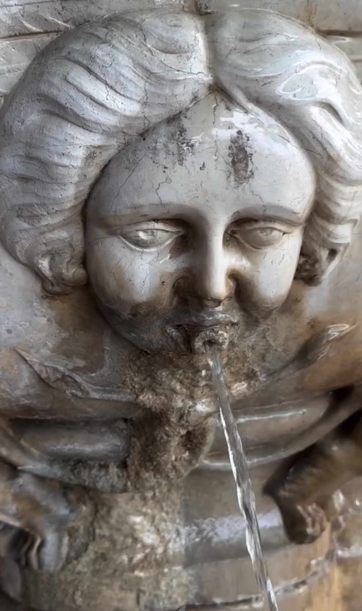 Close-up of a stone fountain with a carved face, water flowing from its mouth, weathered surface, and detailed features.