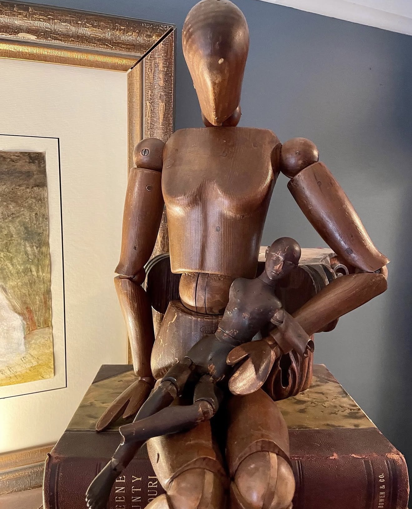 Wooden artist mannequin holding a small black object, standing on a surface with a framed picture in the background.