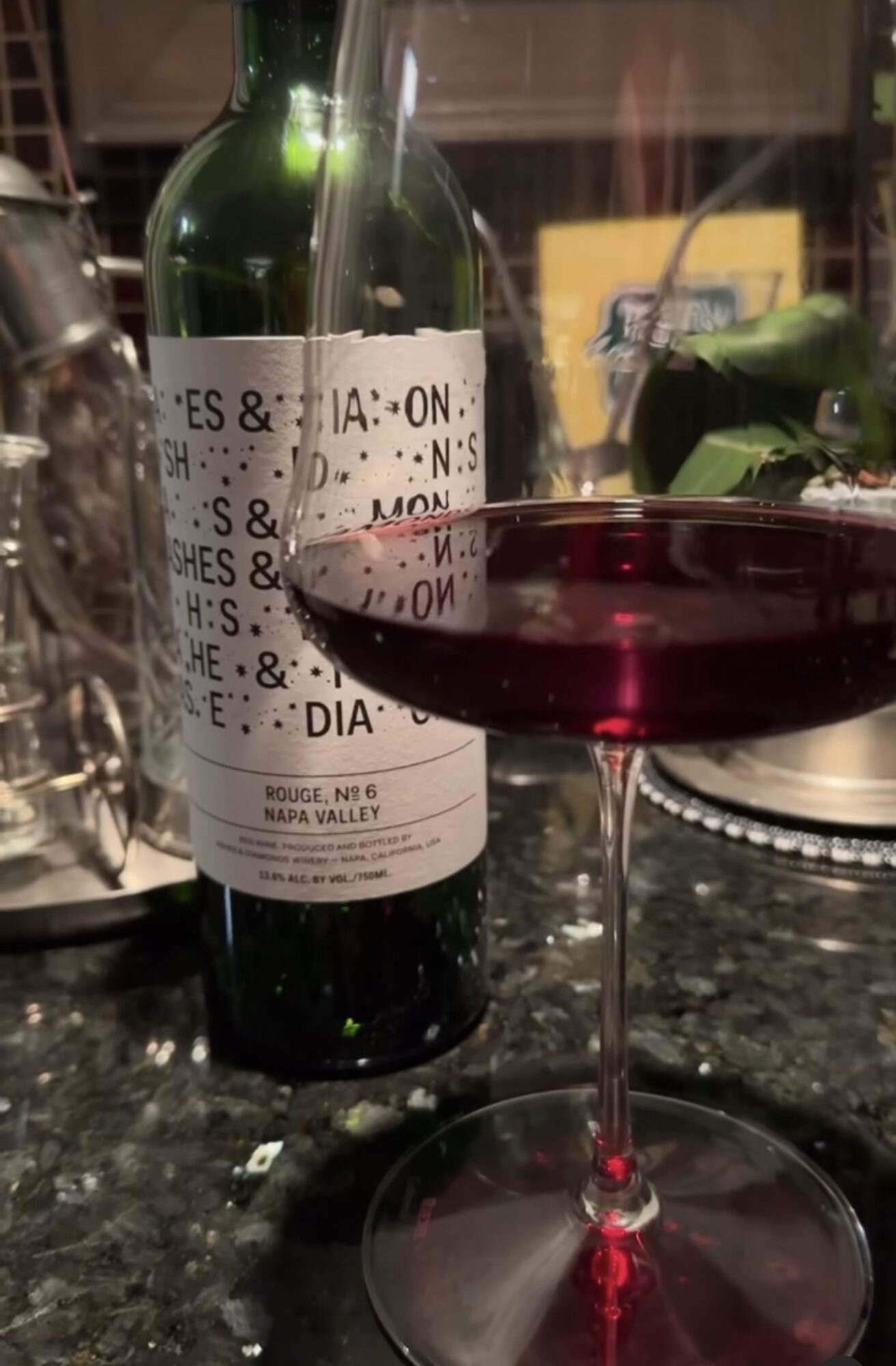 Bottle of wine and a filled wine glass on a table, background shows a window and some objects.