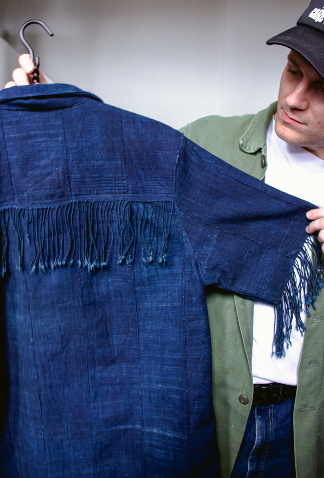 Person holding a blue fringed garment on a hanger, looking at it closely, wearing a cap and green jacket.