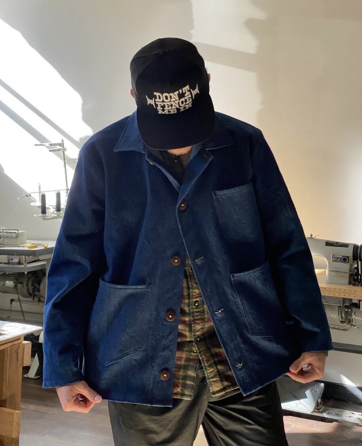 Person wearing a black cap and blue jacket, looking down, in a room with sewing machines and fabric.
