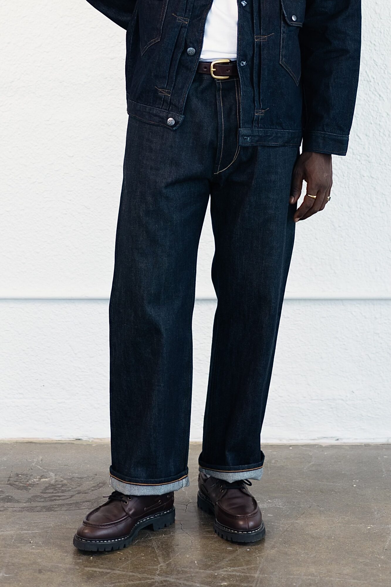 Person wearing dark jeans, a denim jacket, white shirt, and brown shoes standing against a white wall.