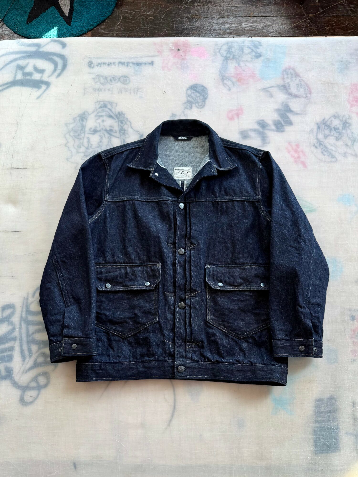 Dark denim jacket with front pockets and button closure, laid flat on a surface with colorful markings