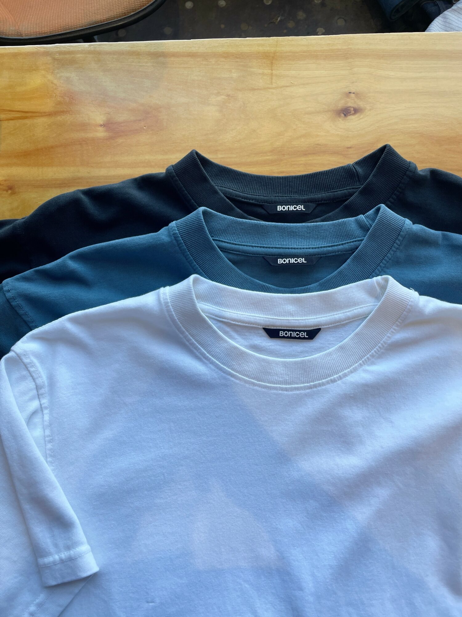 Three folded t-shirts in black, blue, and white on a wooden surface.