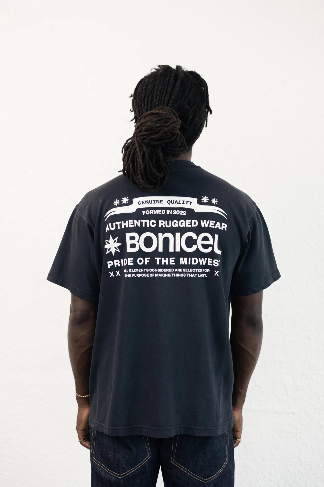 Person with dreadlocks wearing a black T-shirt with white text, facing away from the camera, against a plain white background.